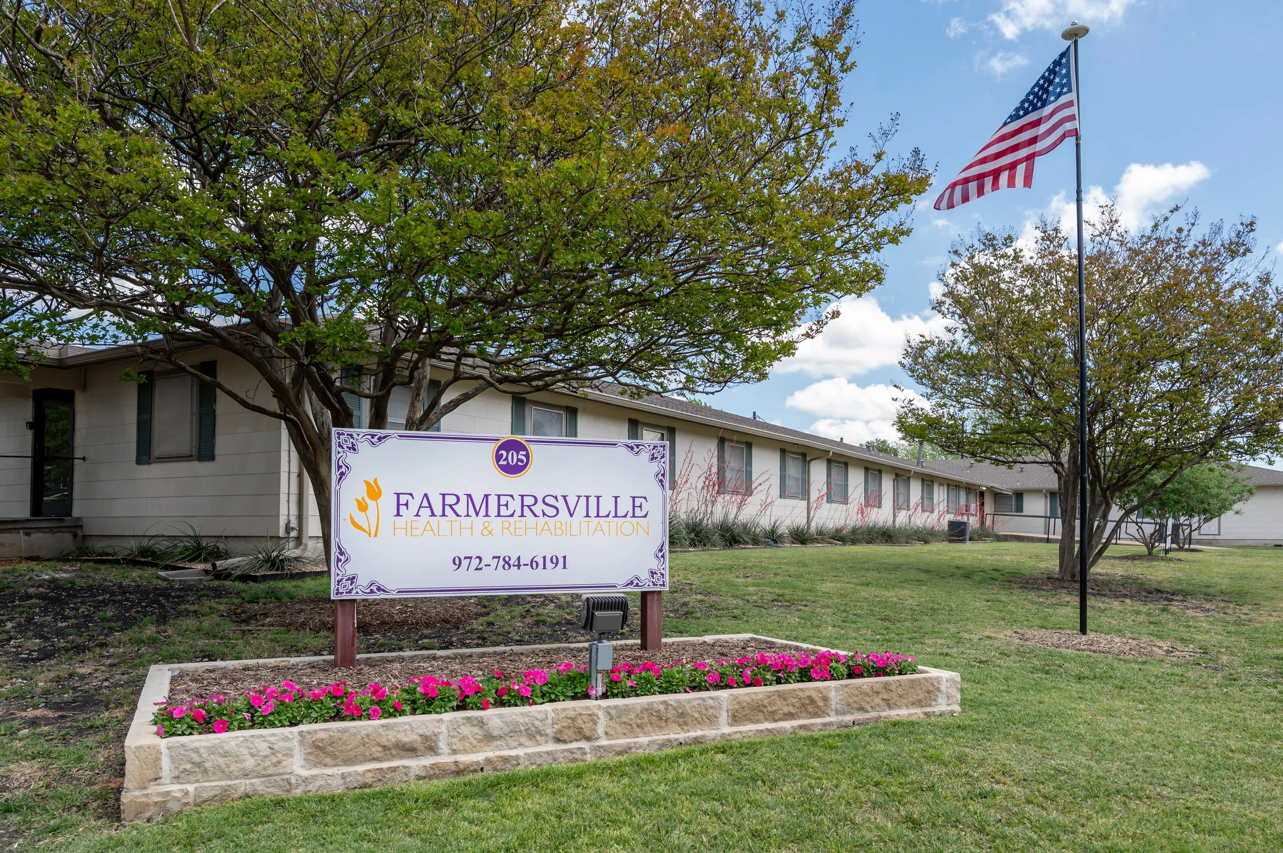 Sign for Farmersville Health & Rehabilitation with phone number, surrounded by a flower bed, trees, and an American flag outside a building under a partly cloudy sky.