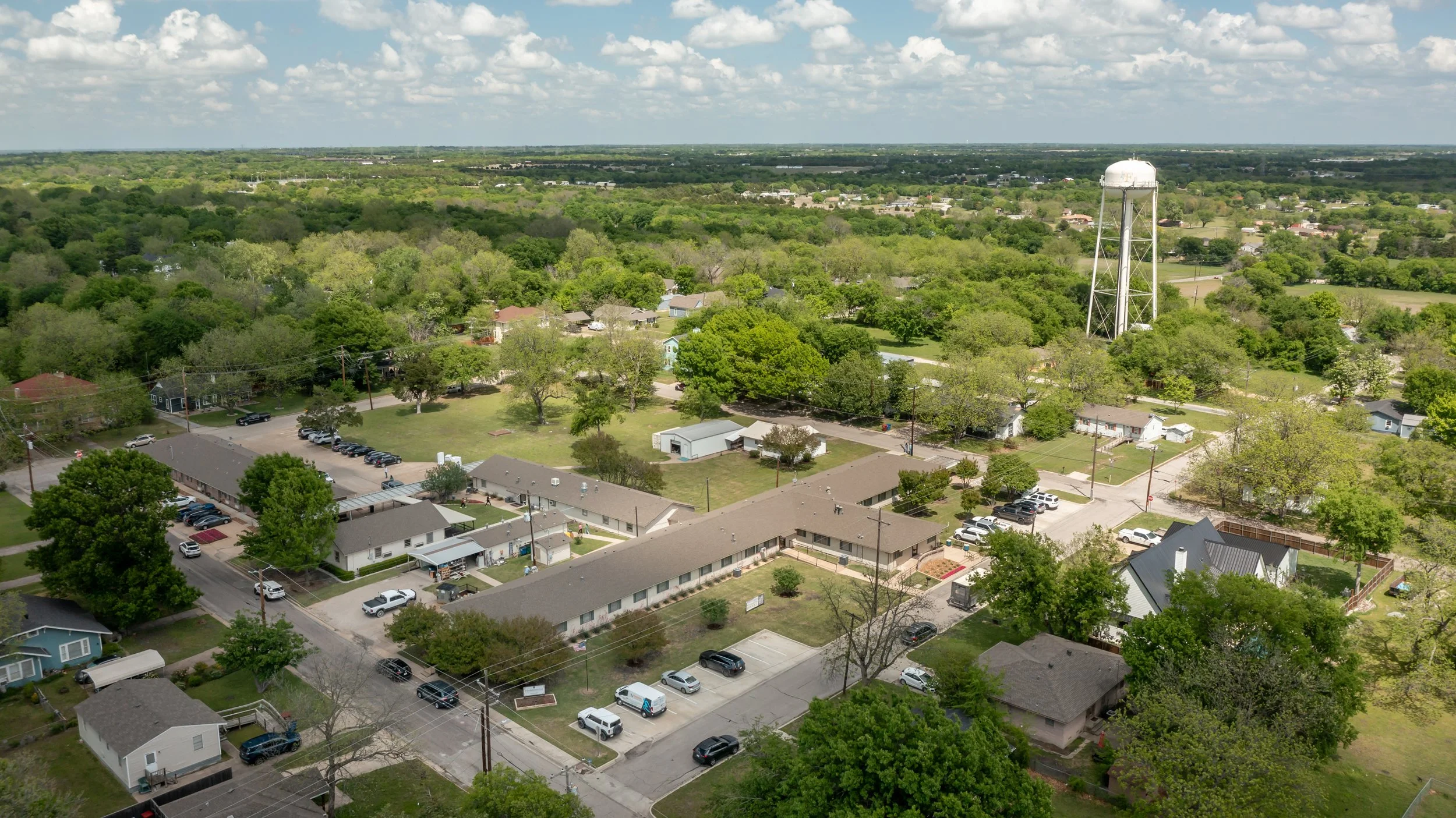Aerial view of a sunny residential neighborhood with multiple single-family homes, trees, and parked cars, and a water tower in the background.
