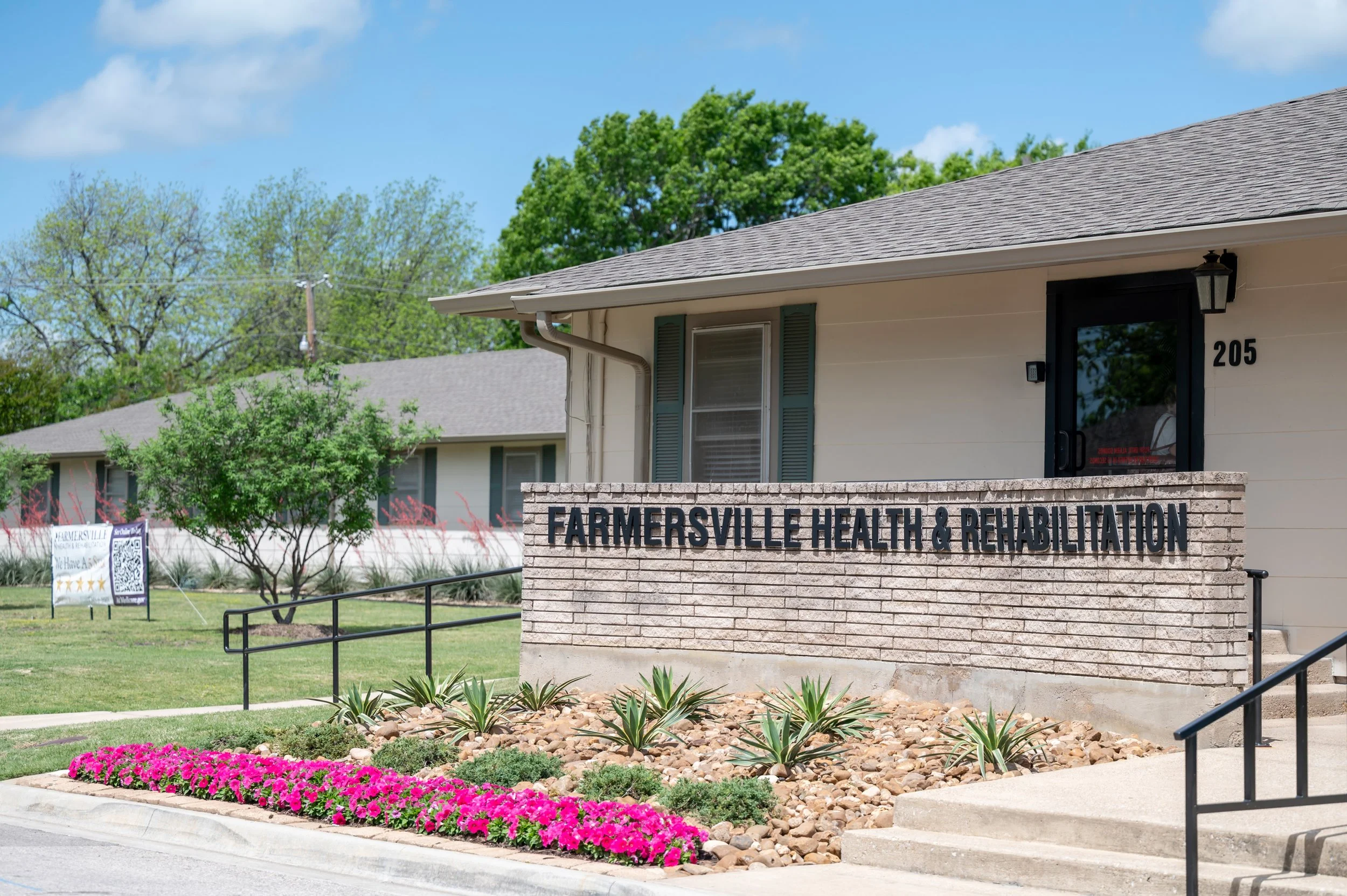 Exterior of Farmersville Health & Rehabilitation building with a brick sign, landscaped flowers, and trees in the background.