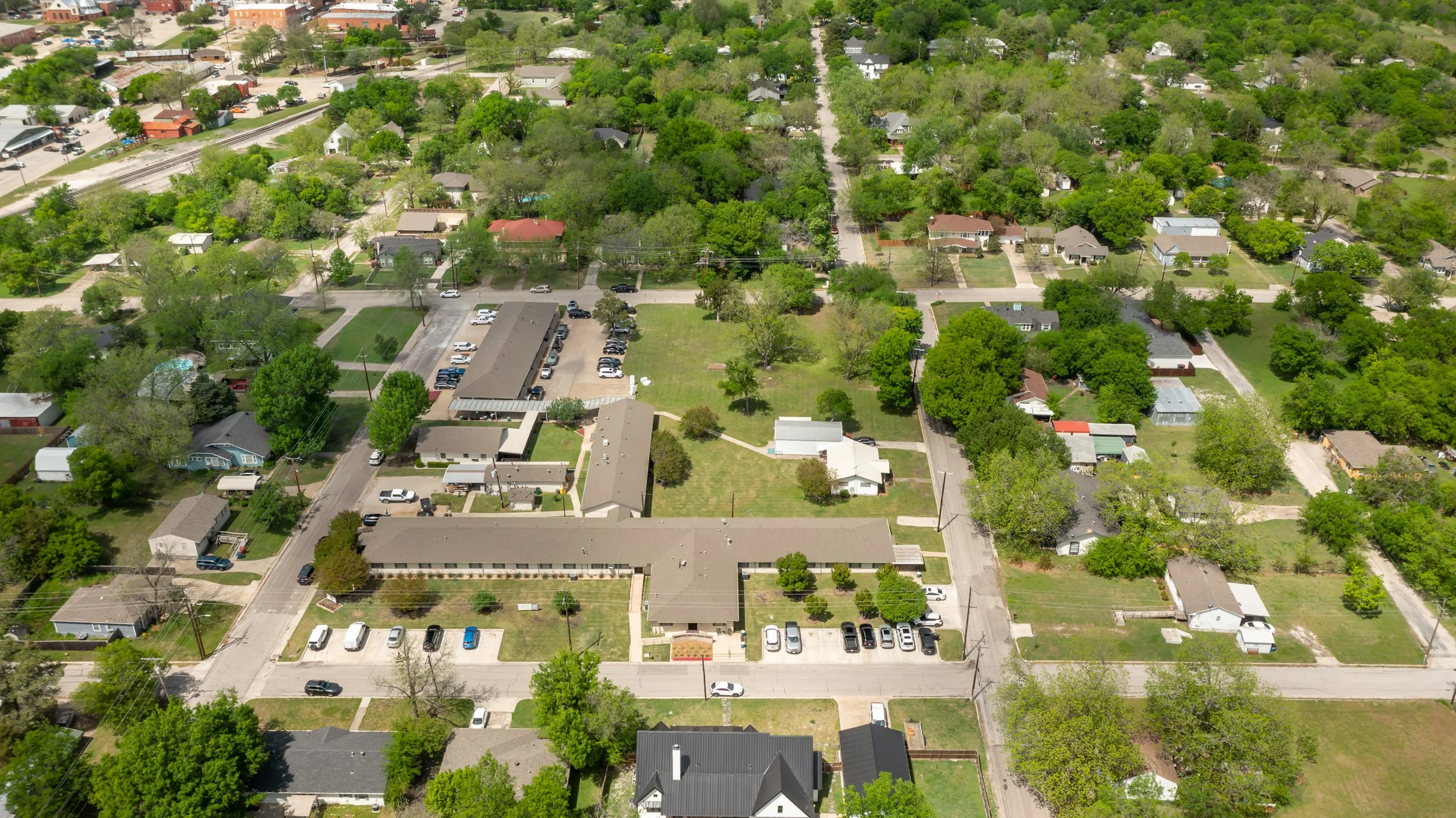 Aerial view of a suburban neighborhood with houses, trees, streets, and parking areas, predominantly green with lush foliage.