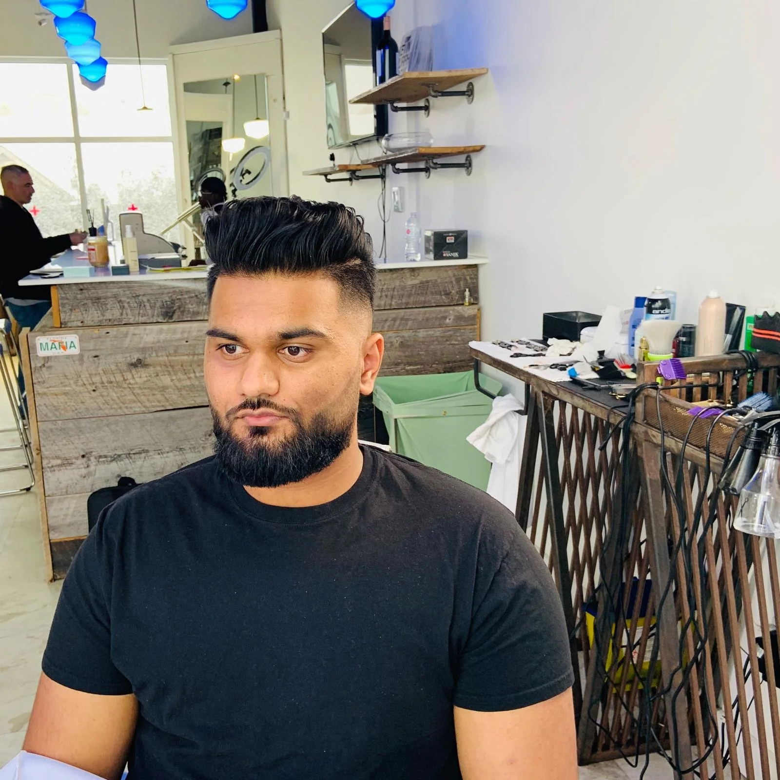 A man with a dark beard and hairstyle sitting in a barbershop or salon. The background features shelves with hair styling tools, products, and miscellaneous items, with another person visible in the distance.