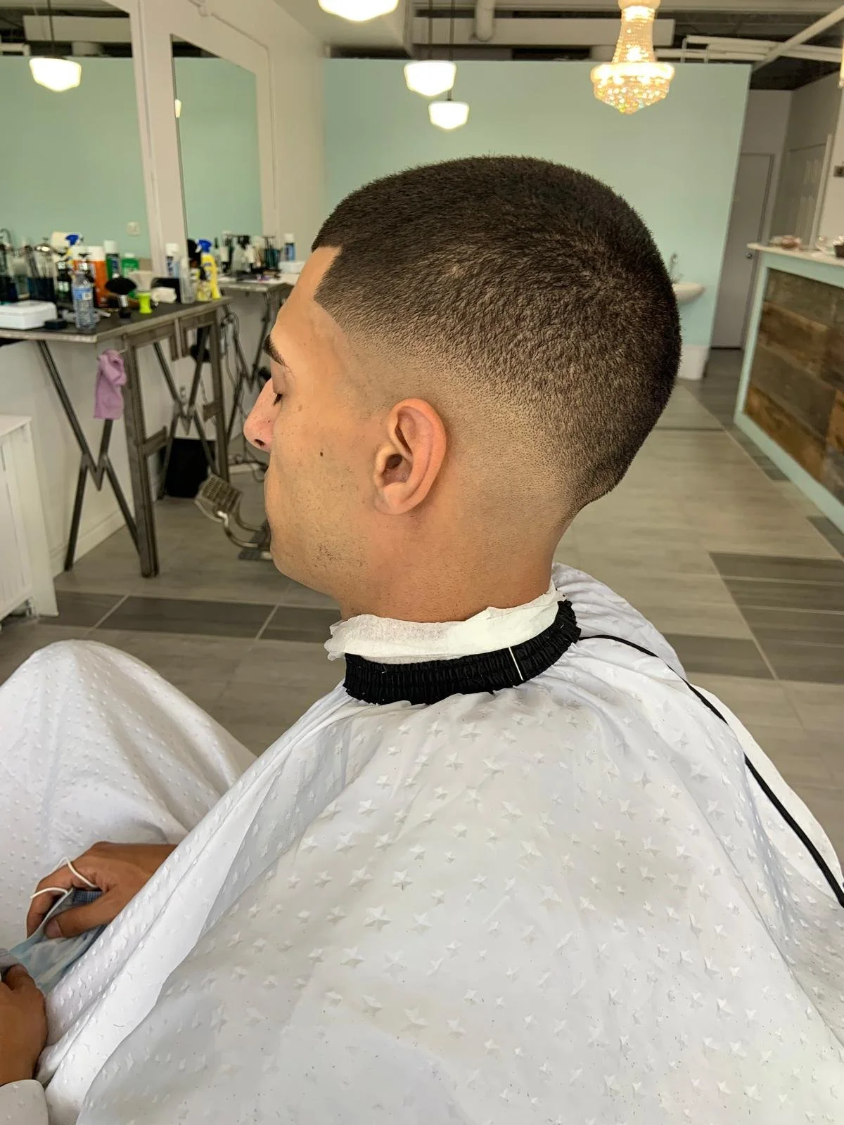 Young man with a freshly cut fade hairstyle sitting in a barber shop or salon, with a barber's cape on, and various hair styling tools and products in the background.