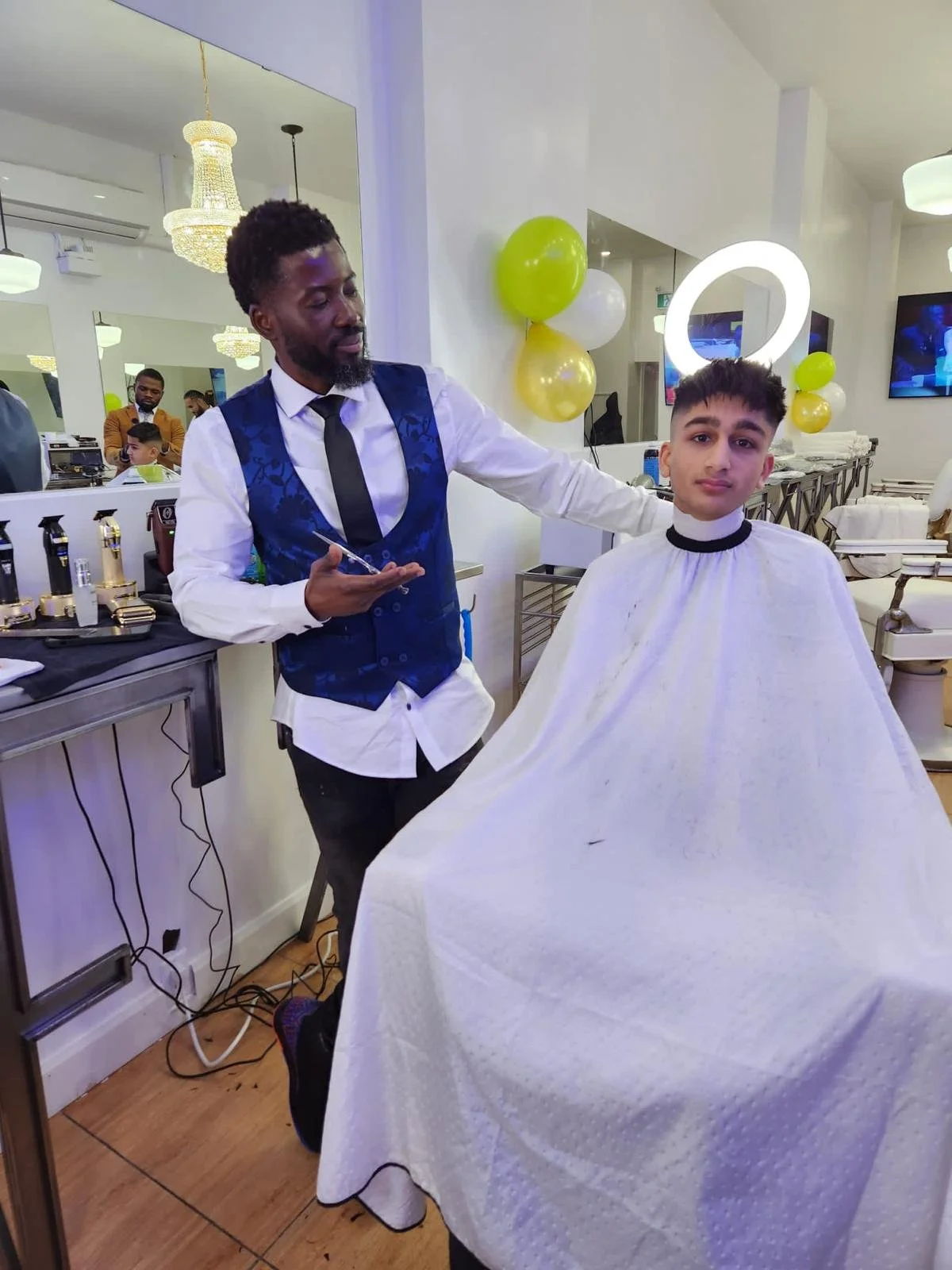 A young man getting a haircut in a salon, with a barber wearing a blue vest and holding hairstyling tools. The salon is decorated with balloons and has mirrors and televisions on the wall.