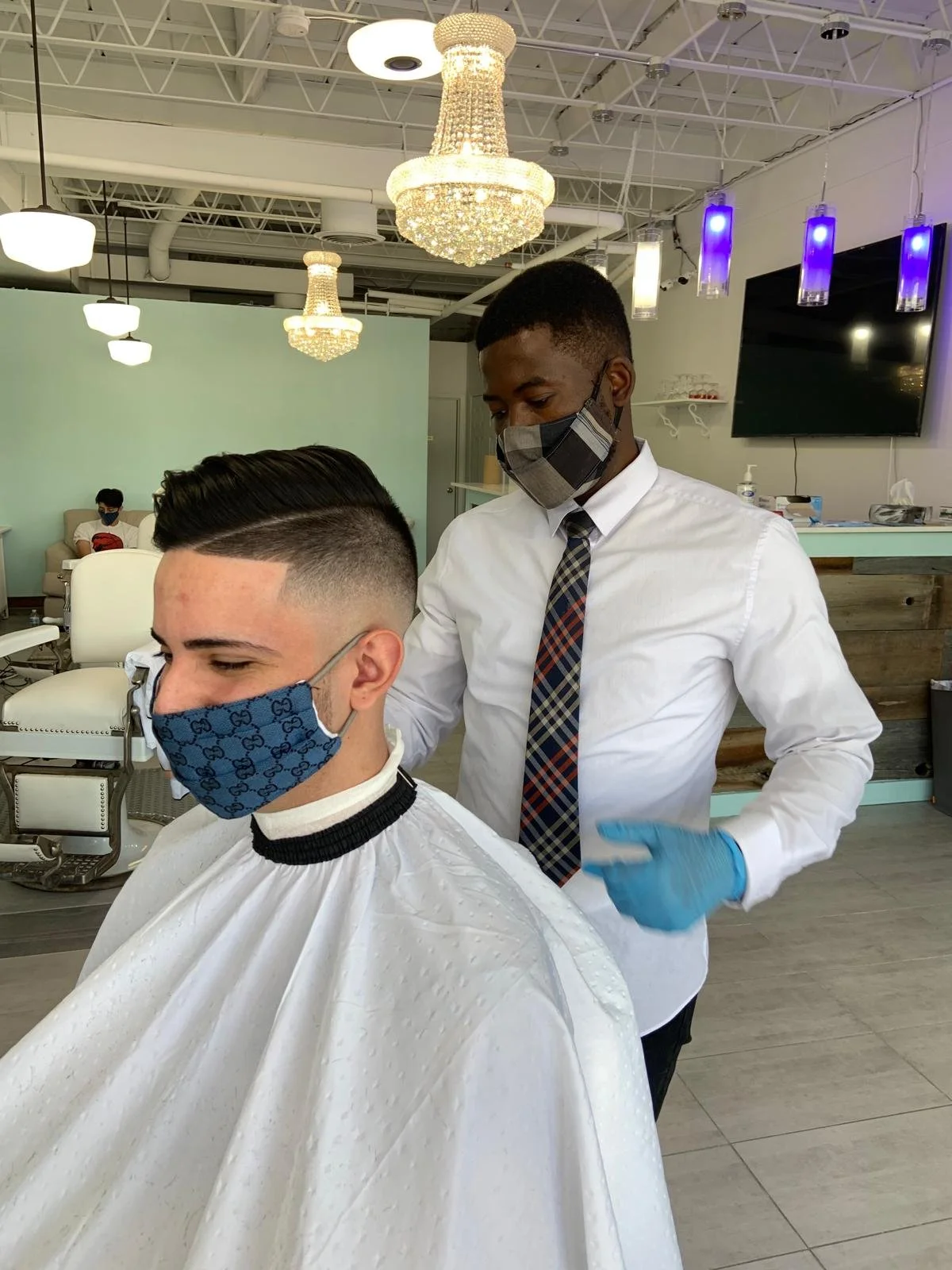 A young man getting a haircut at a salon. The barber is wearing a plaid face mask, gloves, a white shirt, and a plaid tie, while the customer is wearing a gucci face mask and a white barber's cape.