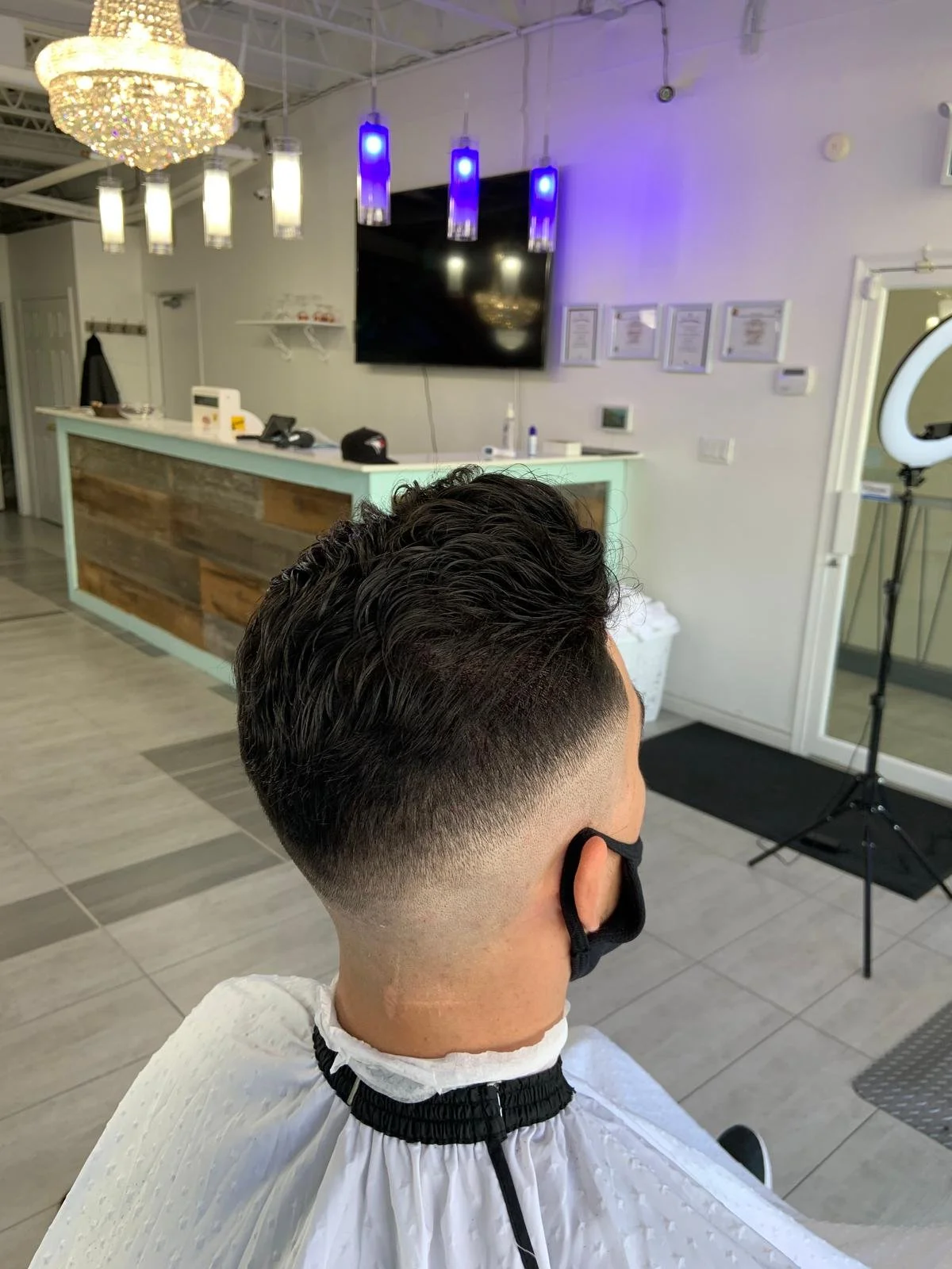 A man with a fresh fade haircut sitting in a barbershop, wearing a white barber cape and a black face mask.