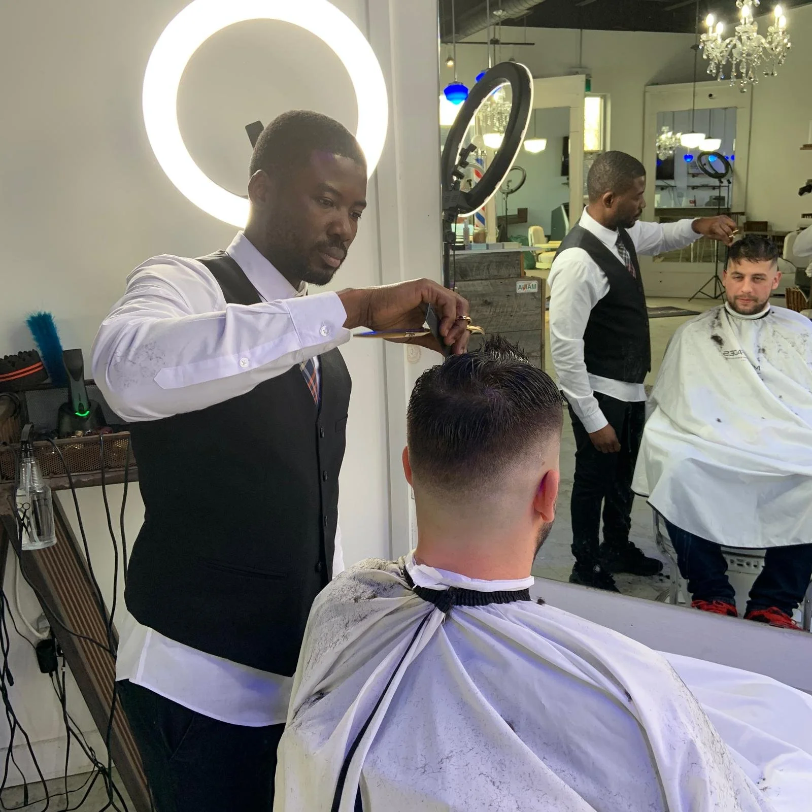 Barber cutting a man's hair in a barbershop. The man is wearing a barber cape. Another barber is visible in the background cutting hair of another customer. The shop has bright lighting, a large mirror, and decorative chandelier.
