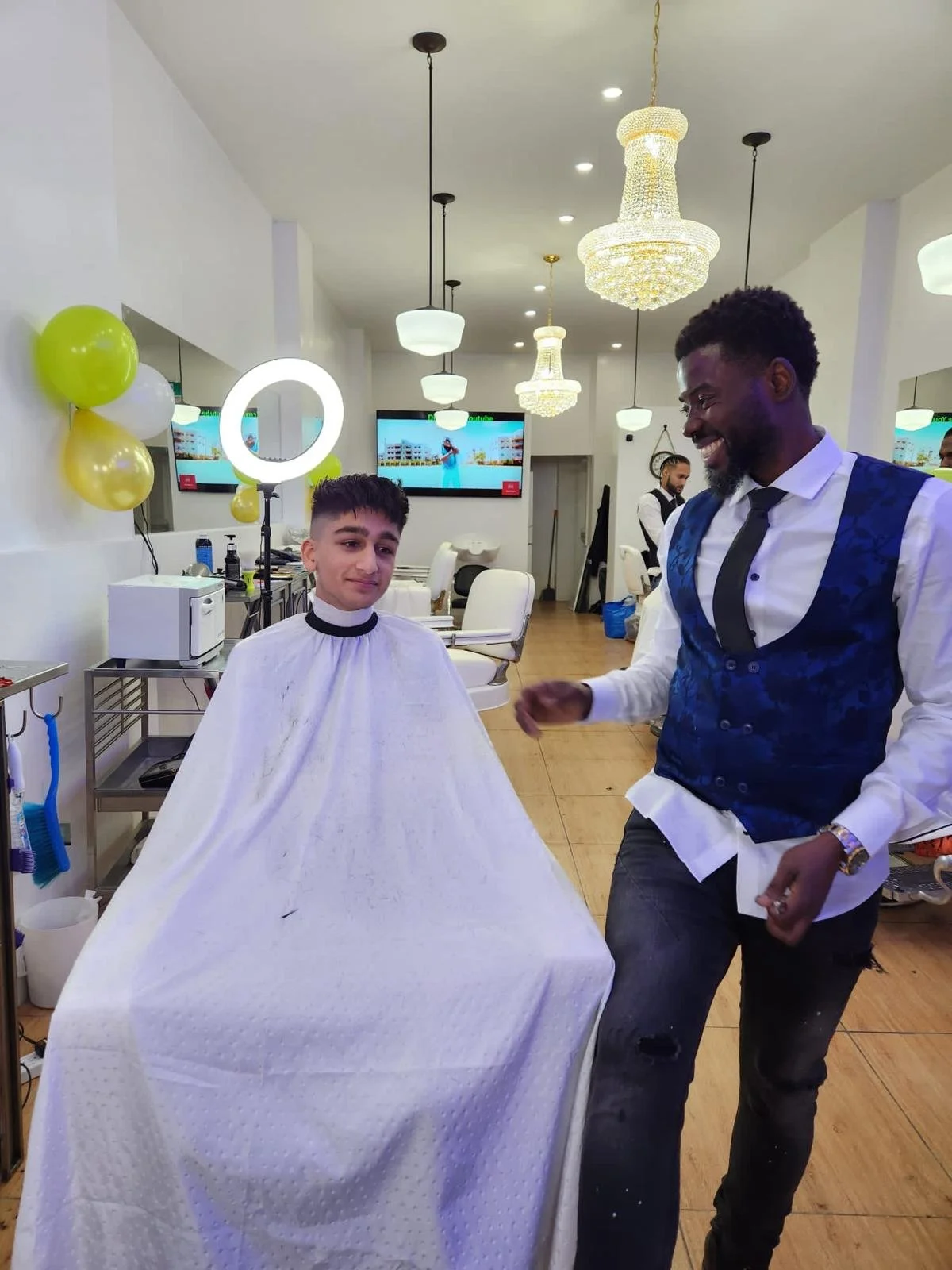 A young boy in a barbershop chair wearing a white cape, smiling while a barber in a vest and tie jokes with him, with balloons and modern lighting in the background.