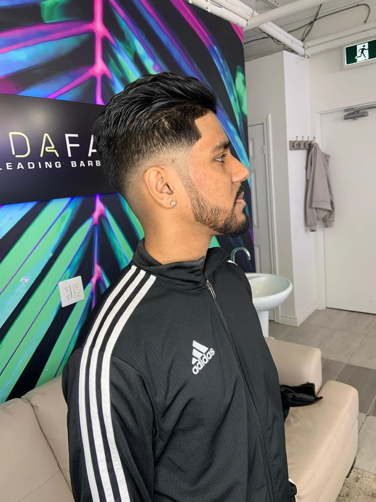 Side profile of a young man with a fresh hairstyle, beard, earrings, wearing a black Adidas sports jacket, sitting on a beige sofa in a barbershop.