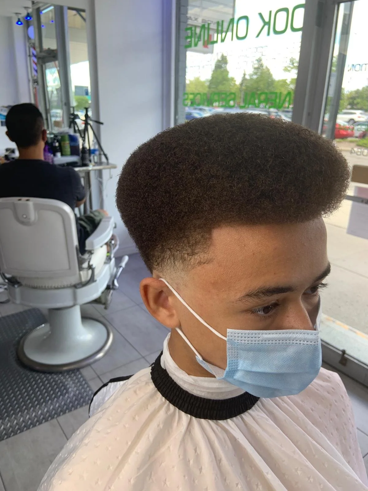 Young man with a fresh hairstyle sitting in a barber shop, wearing a face mask and a barber cape, with another customer and barber tools visible in the background, and a large window showing outside parking lot.