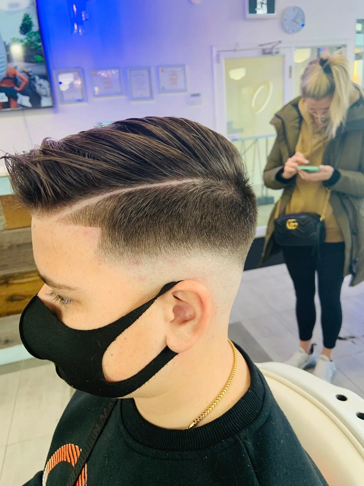 A young man with a fresh haircut featuring a side part and a fade, wearing a black face mask, black shirt, and a gold chain. A woman in the background is looking at her phone inside a barbershop or salon.