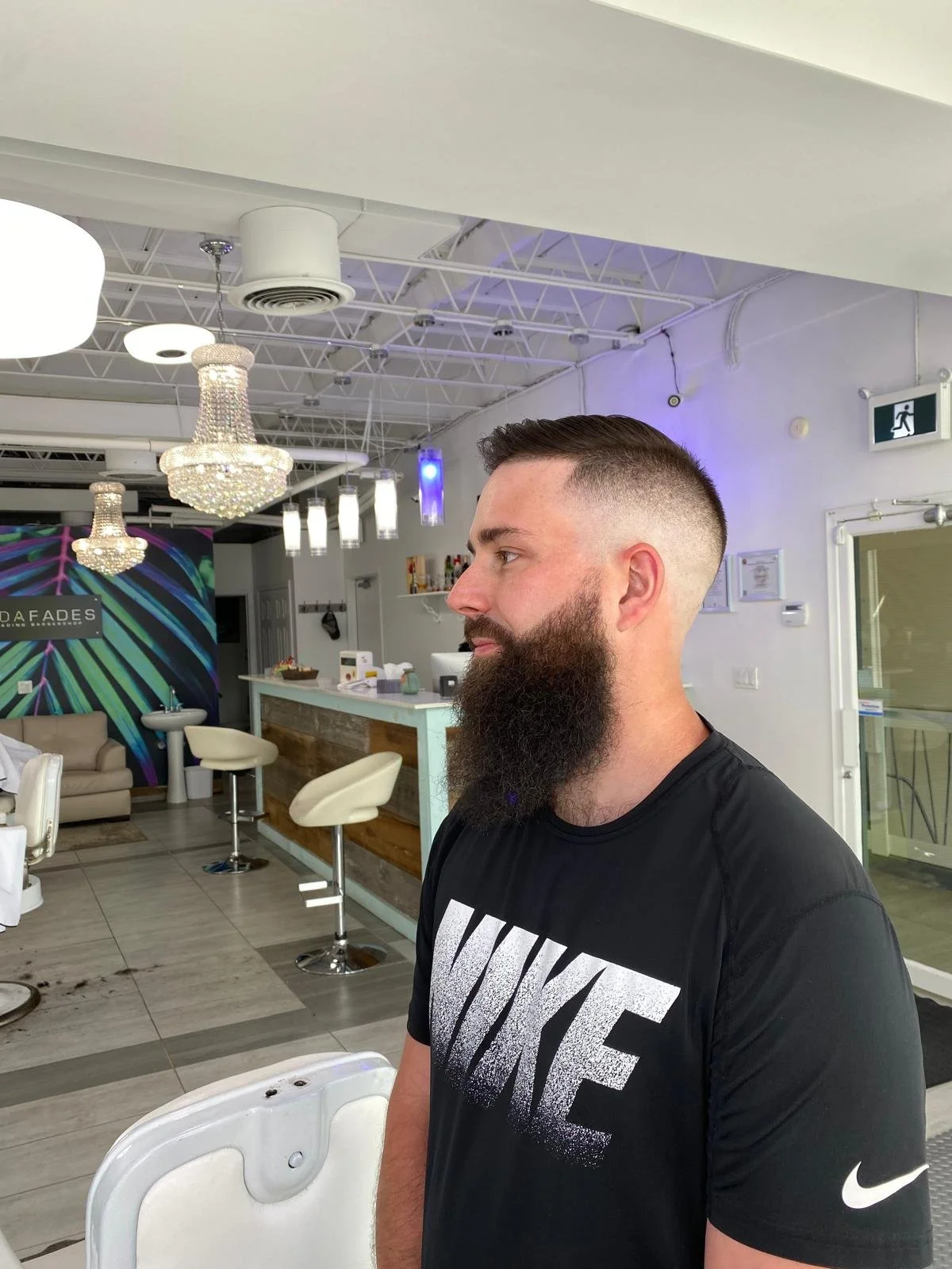 A man with a beard and short haircut, wearing a black Nike t-shirt, standing inside a salon or barbershop with modern decor, chandeliers, and a reception counter.