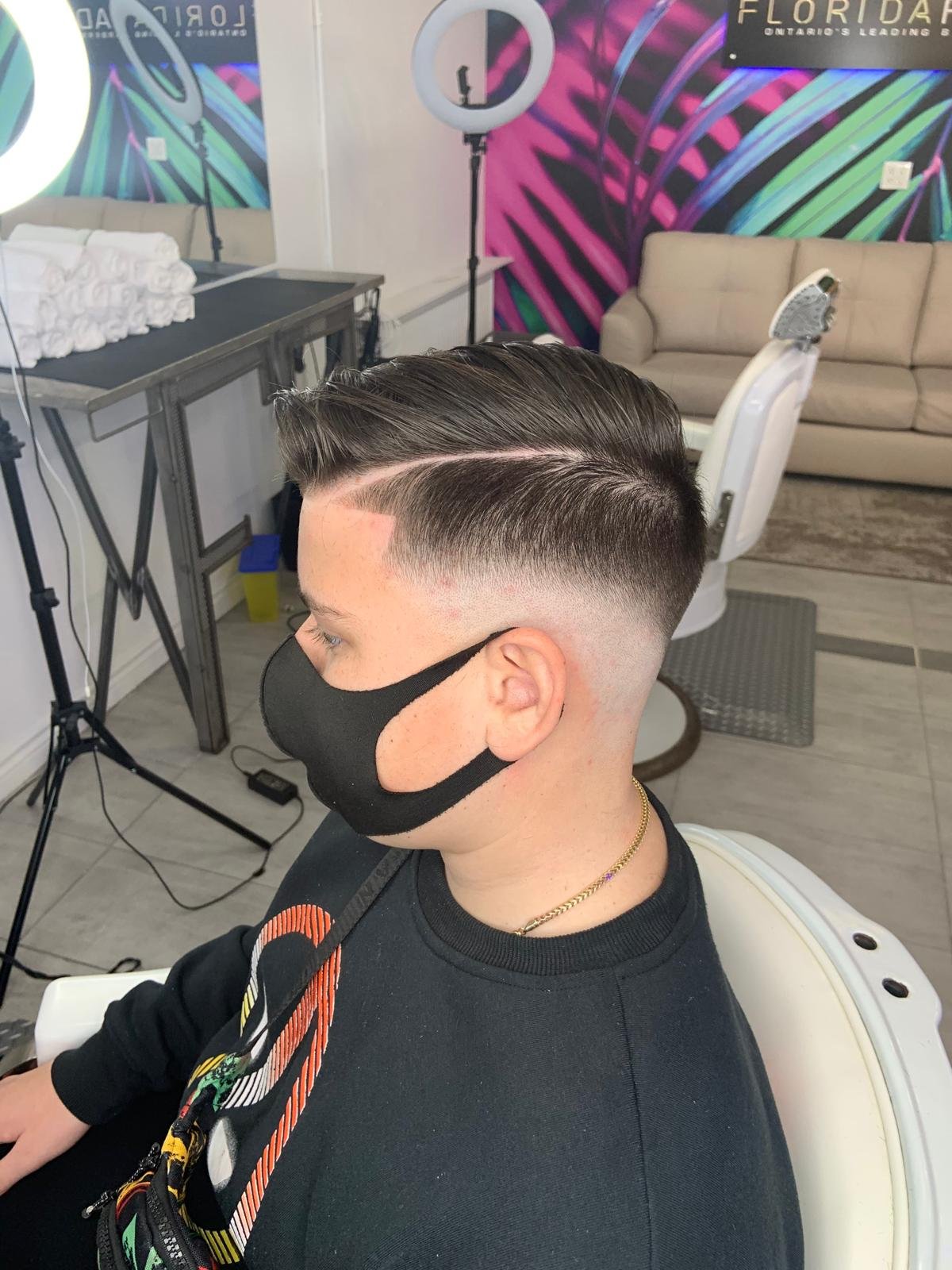 Young man with a freshly cut hairstyle, side profile, wearing a black face mask and black shirt, sitting in a salon with colorful mural walls, bright ring light, and salon equipment in the background.