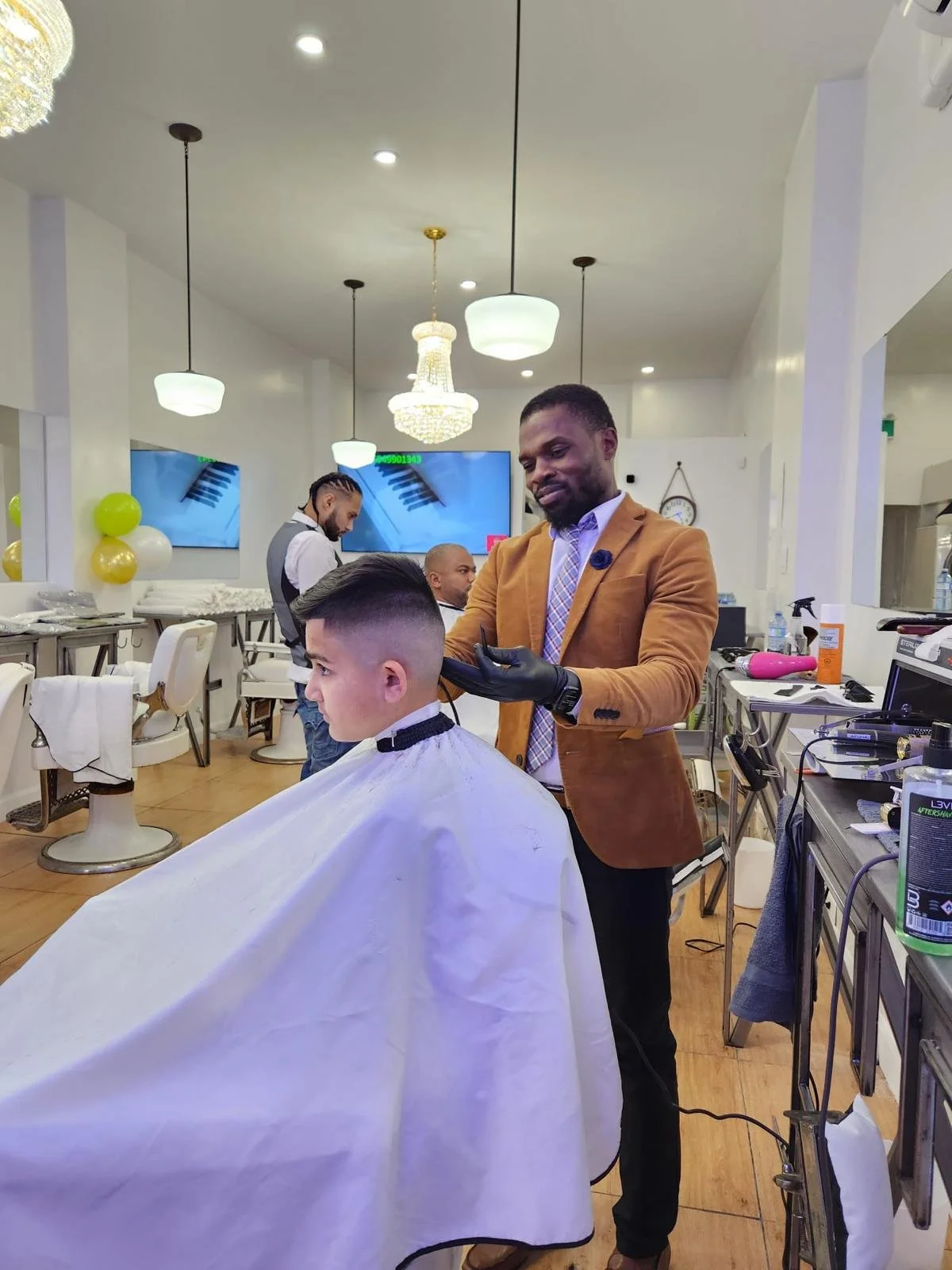 A man giving a young boy a haircut in a salon. The man is wearing a brown jacket and gloves, and the boy is seated with a white cape around his shoulders. The salon is decorated with balloons and has a large screen on the wall, with other people and 