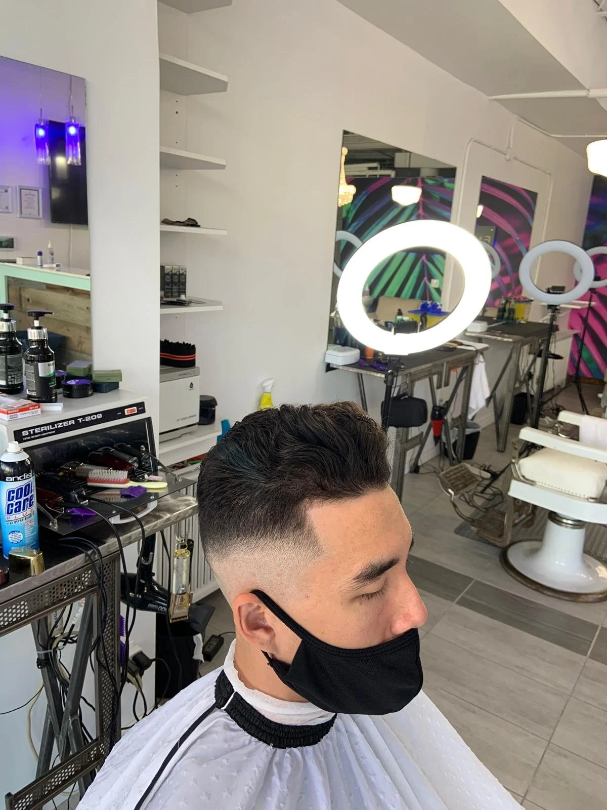 Young man with a fresh fade haircut sitting in a barbershop, wearing a black face mask and a white cape, with modern salon equipment and bright ring lights in the background.