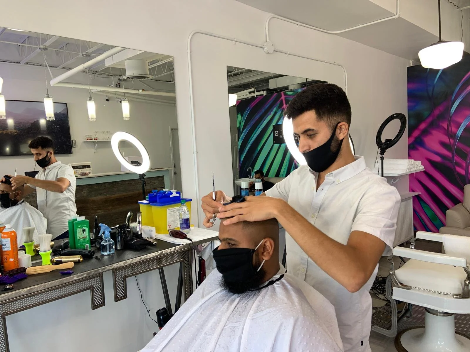 Barbershop scene with barber cutting a customer's hair, both wearing black masks, in a modern salon with mirrors, colorful wall art, and salon tools on the counter.
