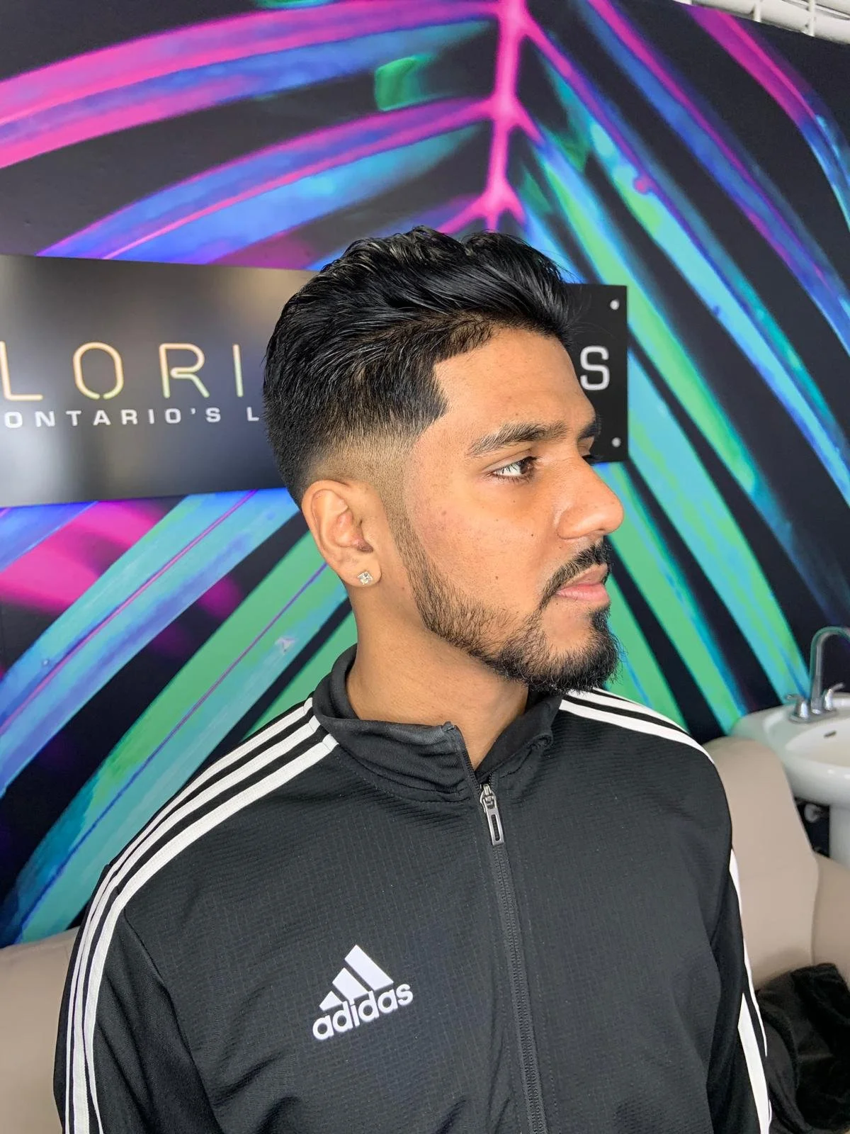 A man with a well-groomed hairstyle, beard, and earrings, wearing a black Adidas sports jacket, sitting in a salon with colorful neon wall art behind him.