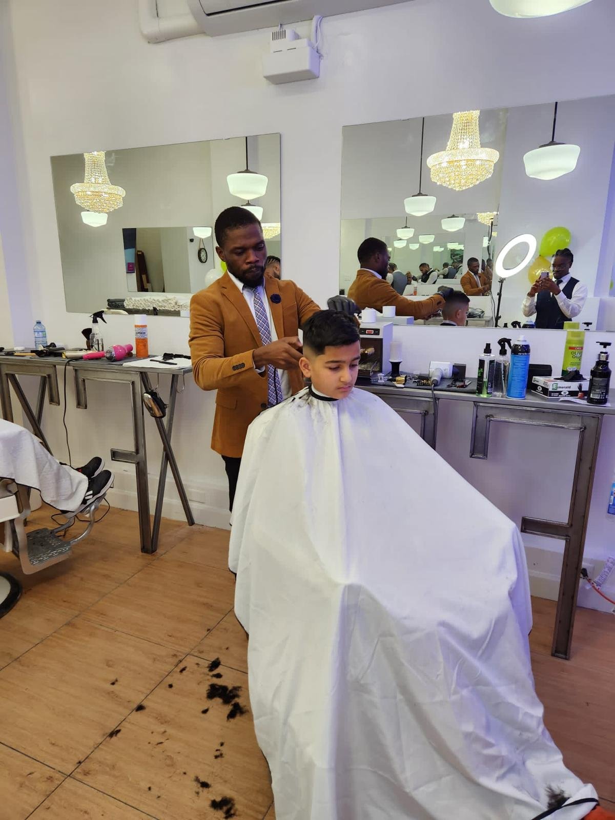 Barber trimming a young boy's hair in a salon with mirrors, chandeliers, and hair styling tools visible.