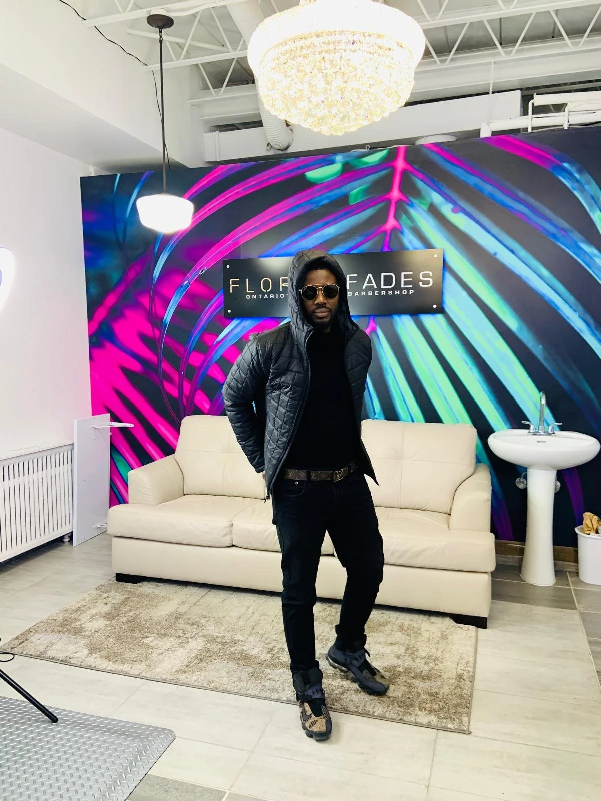 A man dressed in black with sunglasses, standing in a salon or barbershop, in front of a colorful, abstract background mural with the sign 'Florida Fades' in Ontario, Canada.