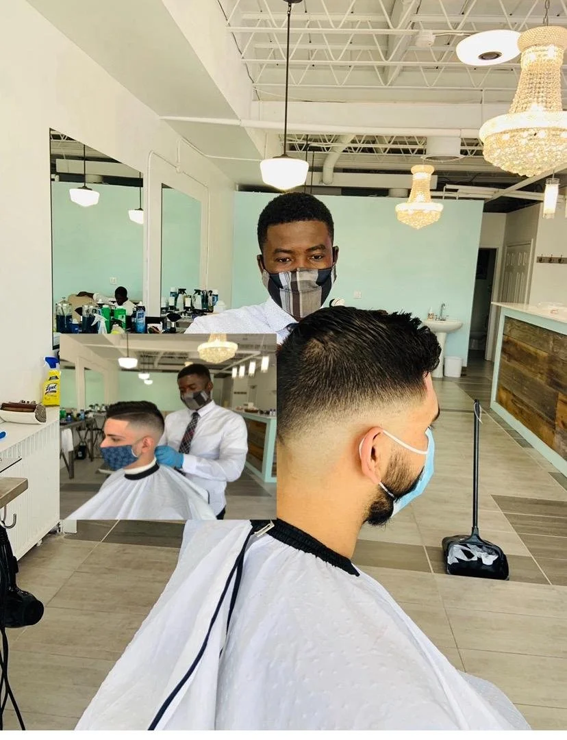 Man in a barber shop getting a haircut, with barber wearing a mask and gloves, and a mirror reflecting his face.