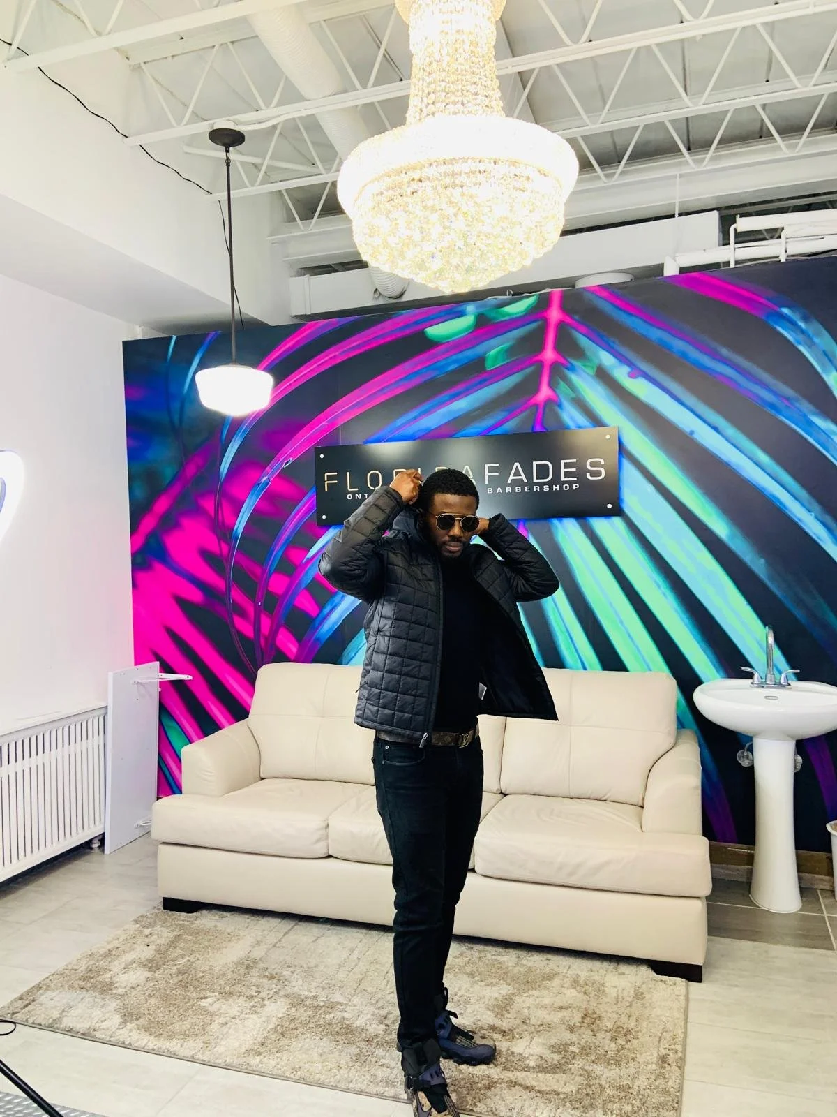 A man wearing sunglasses and a black quilted jacket adjusting his hair in front of a modern colorful neon wall at a barbershop named 'Flow Fades'. He is standing on a small rug in front of a white sofa, with a sink visible to his right and a chandeli