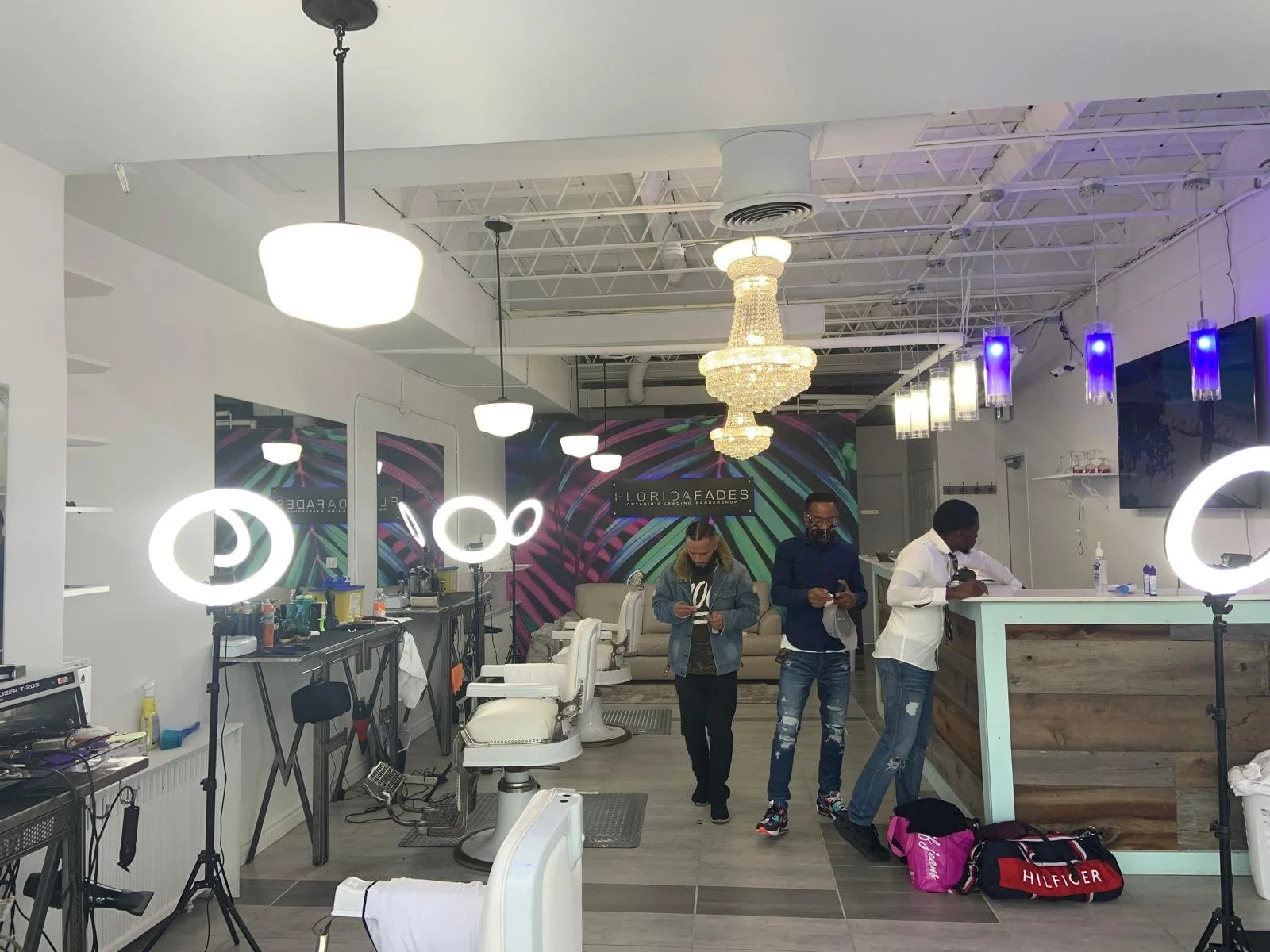 Interior of a modern salon or barbershop with three men standing at the reception counter, working or ordering. The salon has white walls, modern lighting fixtures, and a decorative chandelier. There are salon chairs, mirrors, and various hairdressin