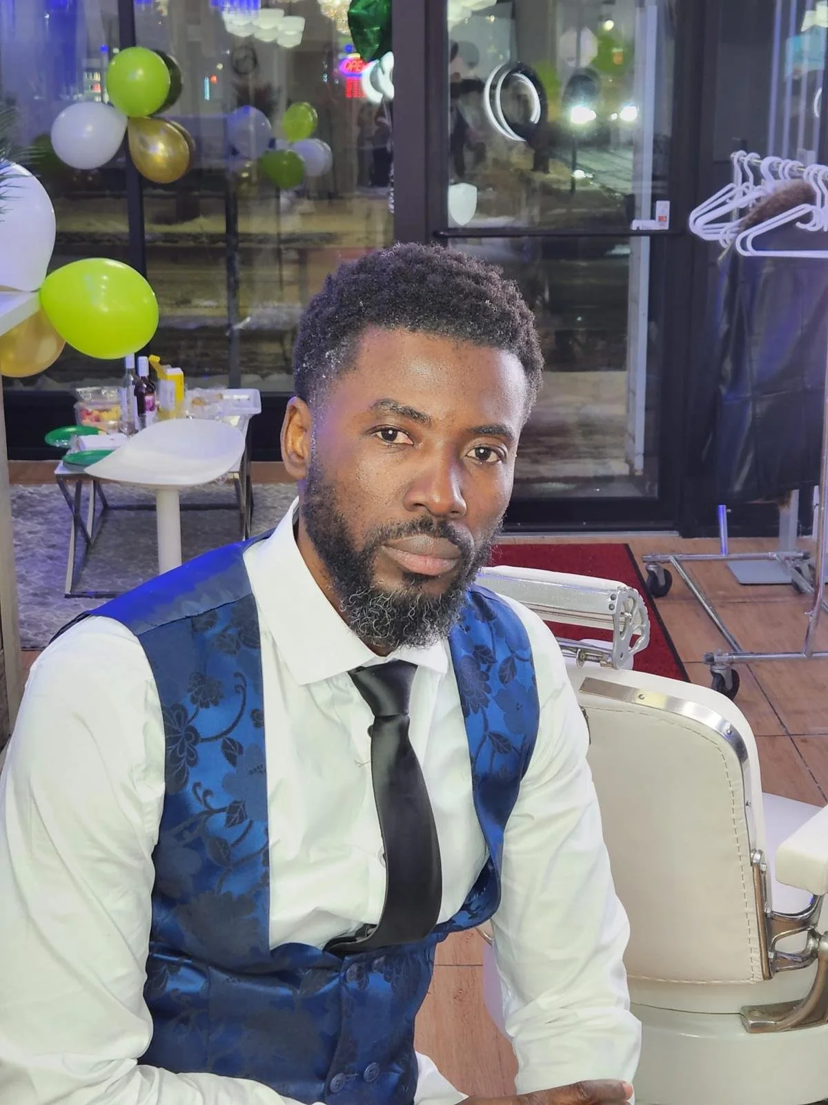 A man with a beard and short curly hair, wearing a white shirt, a black tie, and a blue vest with floral patterns, sitting indoors near a window decorated with green, white, and gold balloons, with a vintage barber chair and coat rack in the backgrou