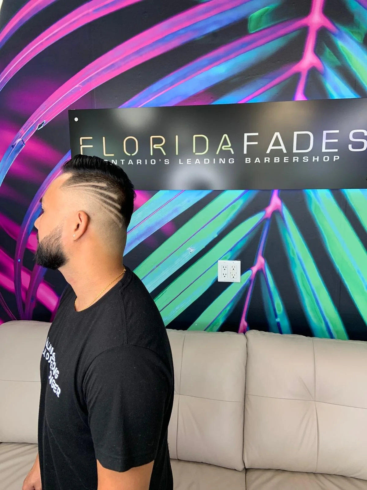 A man with a roller hairstyle and beard sitting on a beige couch in front of a vibrant neon-colored wall with a sign that reads 'Florida Fades: Ontario's Leading Barbershop'.