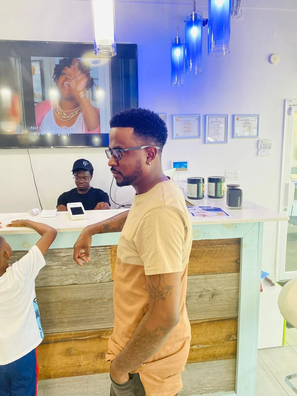A man with glasses and tattoos on his arm standing at a counter in a modern cafe or restaurant, with a boy next to him. In the background, a woman is on a TV screen with subtitles.