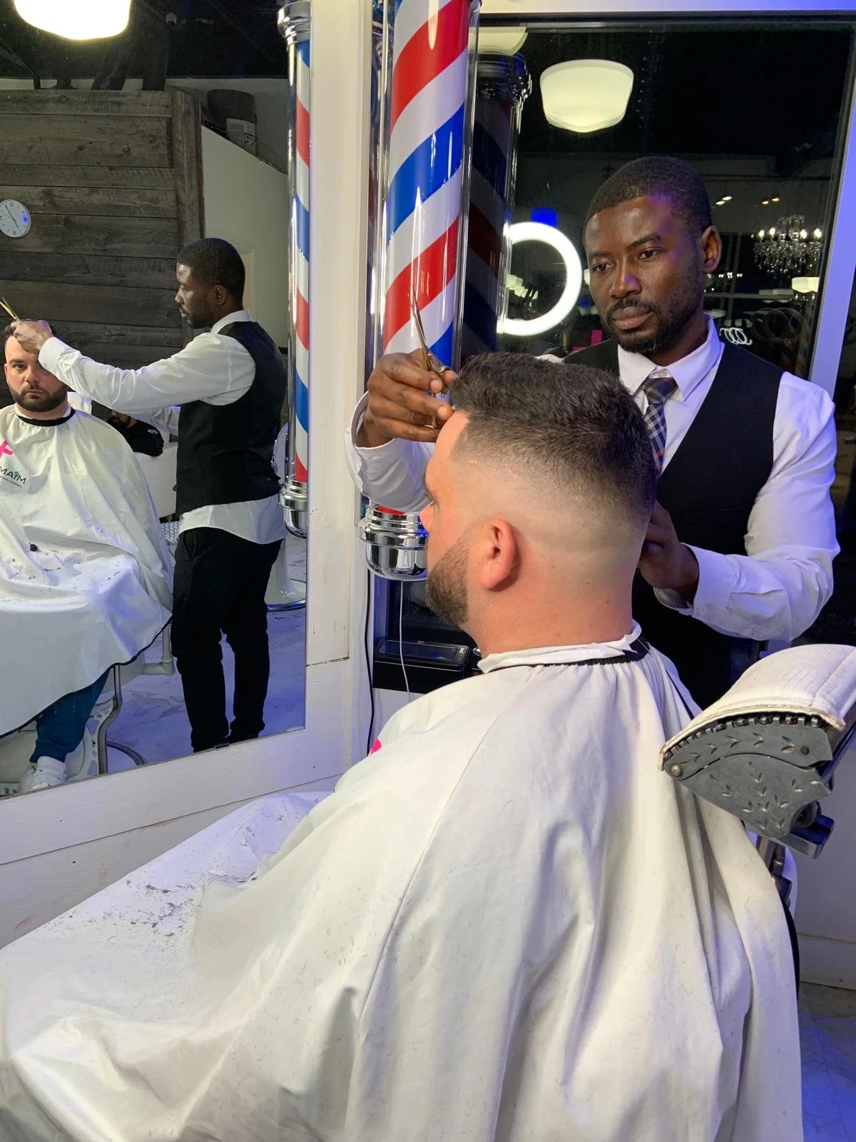Barber giving a male client a haircut in a barbershop. The barber, dressed in a white shirt and black vest, is trimming the client's hair, who is seated in a barber chair with a cape.