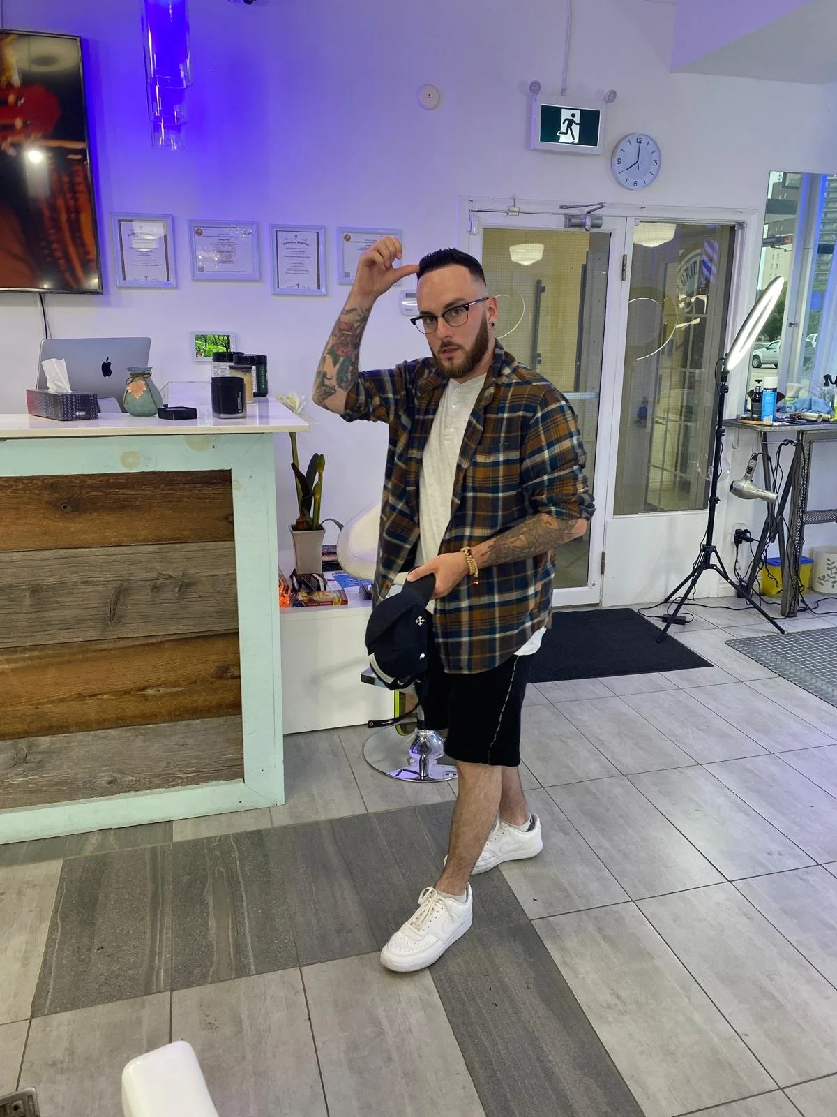 A man with glasses, a beard, and tattoos, wearing a plaid shirt, shorts, and white sneakers, standing indoors with one hand on his head and holding a black cap in the other hand.