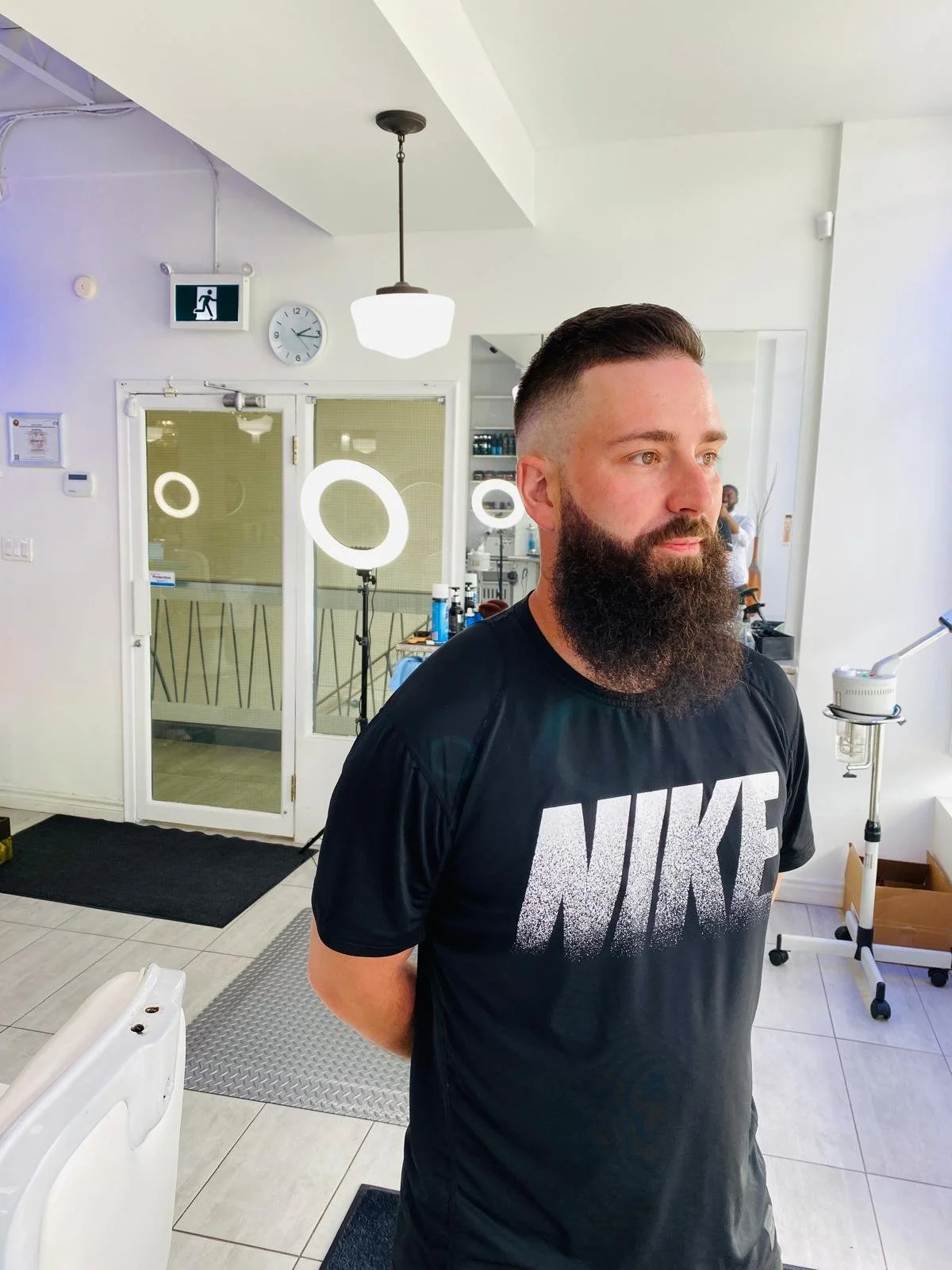 A man with a thick beard and short hair, wearing a black Nike t-shirt, is standing inside a modern establishment, possibly a salon or barbershop, with his arms behind his back. In the background, there are circular ring lights, a door with glass pane