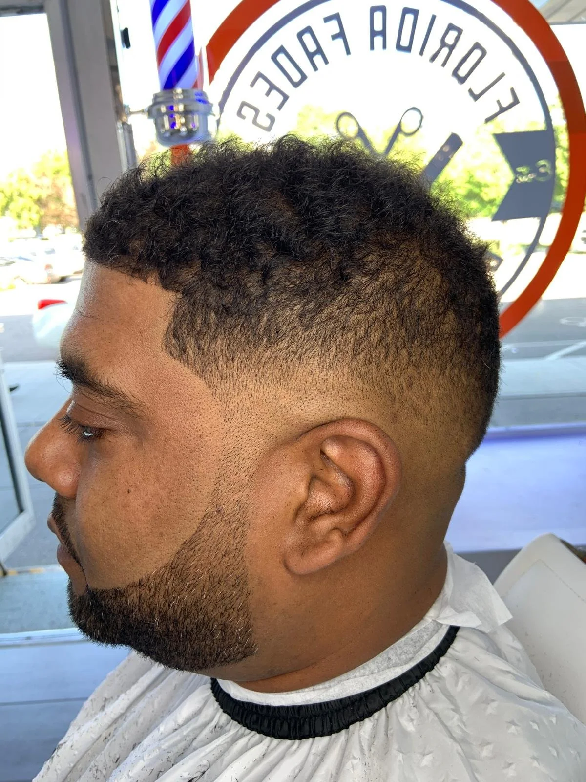Side view of a man with a fresh, high fade haircut with curly hair on top and a beard, sitting in a barber's chair inside a barbershop, with a mirror and a 