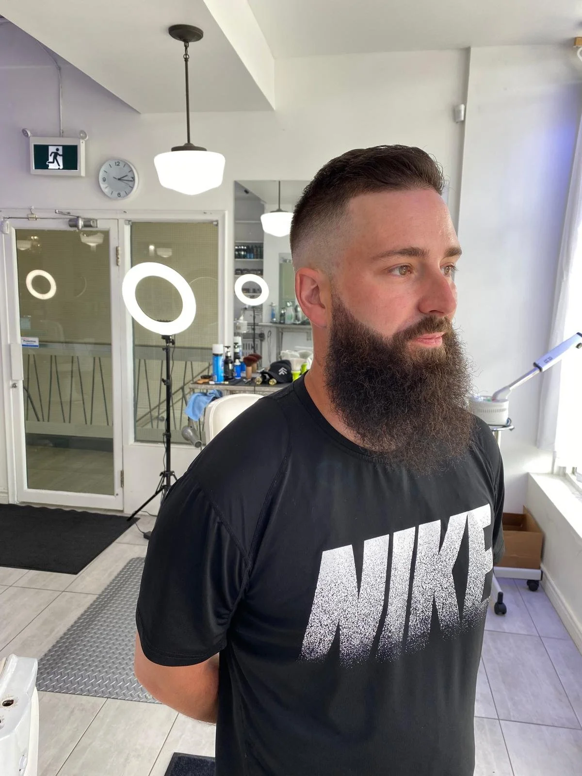 A man with a full beard and short hair standing in a barbershop or salon, wearing a black Nike T-shirt, with mirrors, lighting fixtures, and barber tools visible in the background.