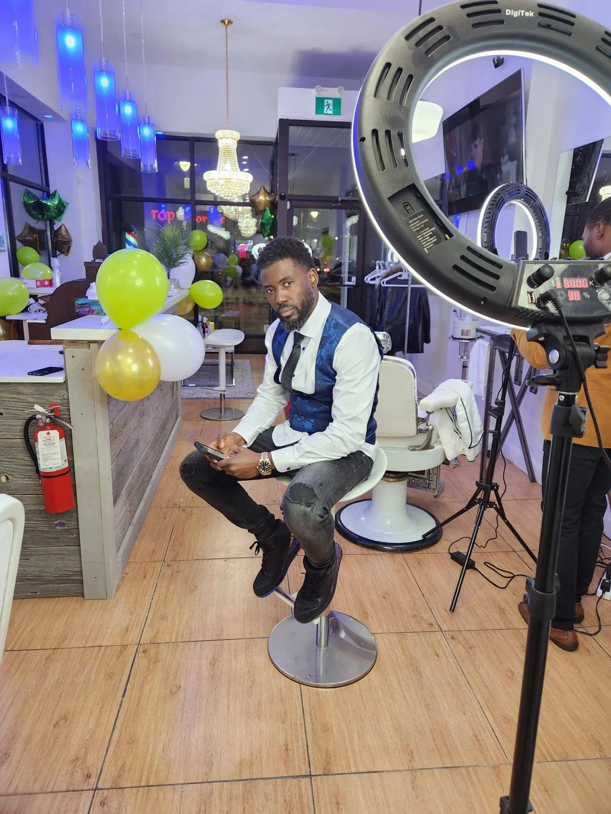 A man with a white shirt and blue vest sitting on a white barber chair in a decorated indoor setting, holding a phone, with a ring light and other equipment visible in the foreground, and balloons and chandeliers in the background.