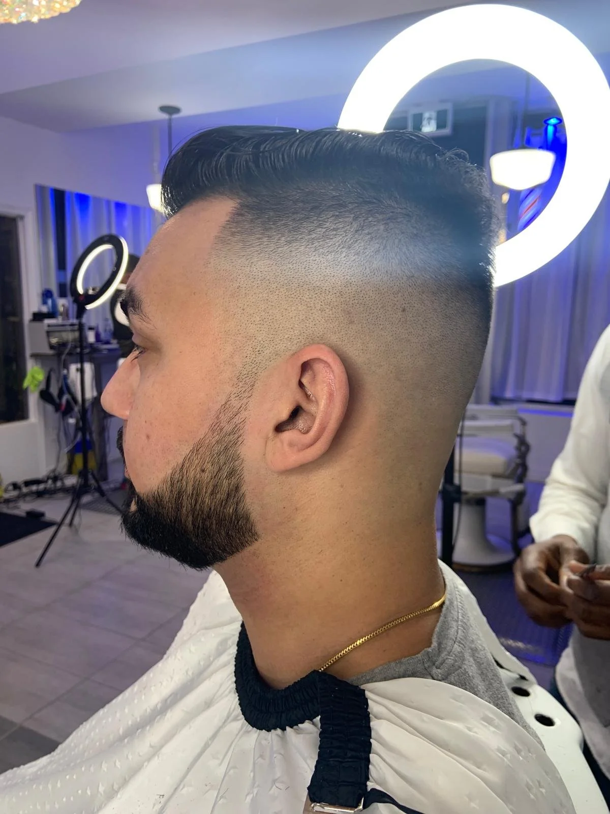 Side view of man with a fresh hairstyle featuring a fade and beard, seated in a barbershop with modern lighting and equipment.
