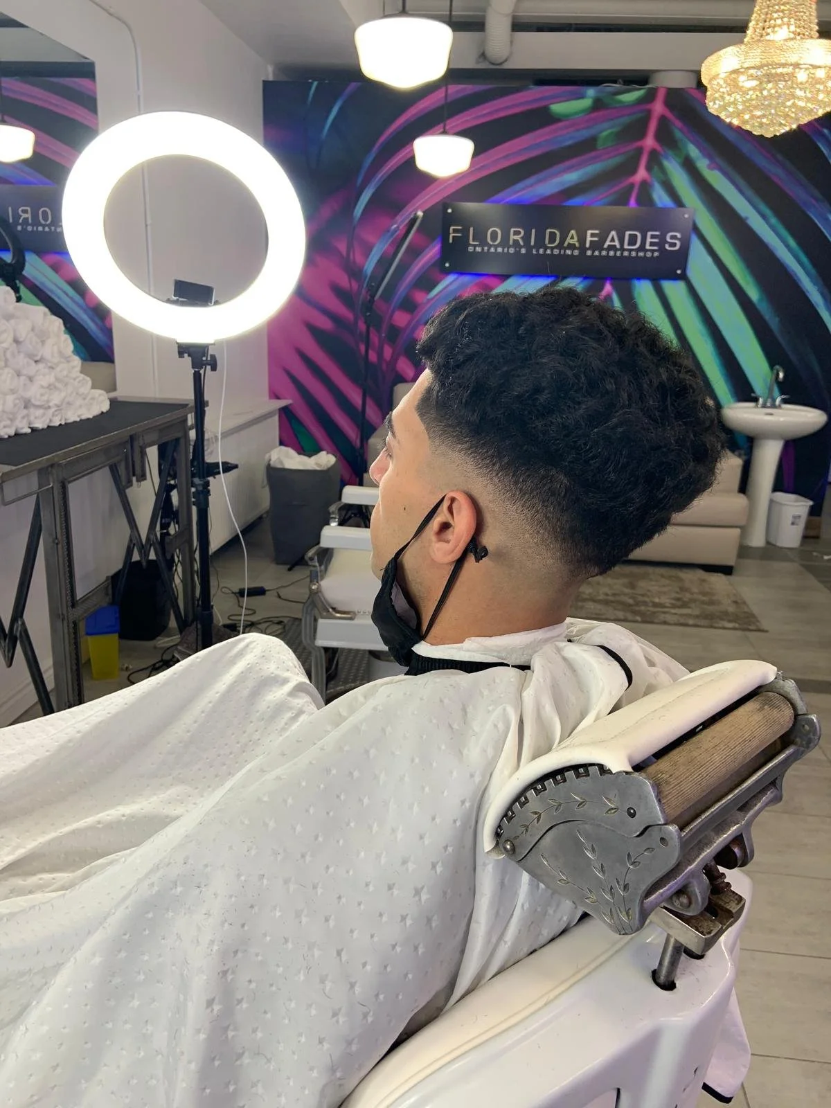 A man with a curly hairstyle sits in a barber chair at a barber shop with a colorful, neon mural background. He wears a face mask and a white barber cape.