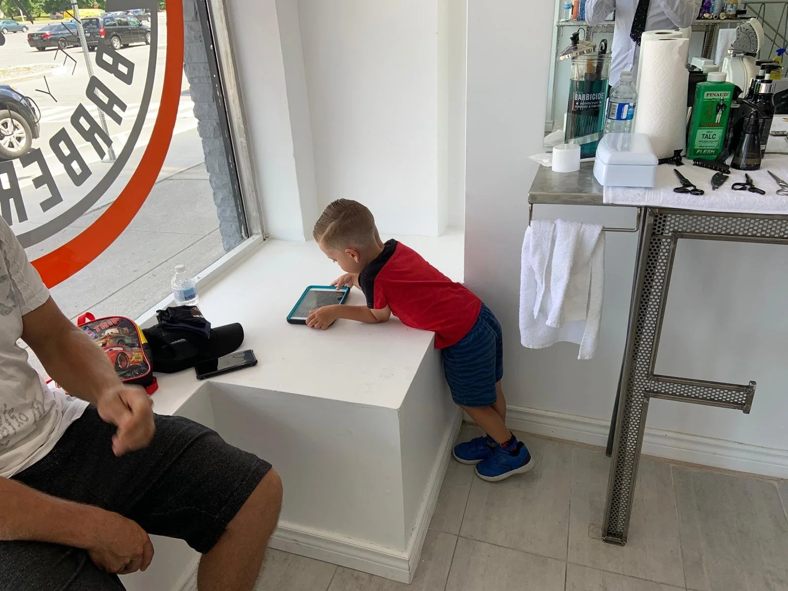 A young boy leans over a windowsill, looking at a tablet screen. He is wearing a red and black shirt, blue shorts, and blue sneakers. An adult sitting nearby has their hand resting on their knee. The scene appears to be inside a tattoo shop, with a w