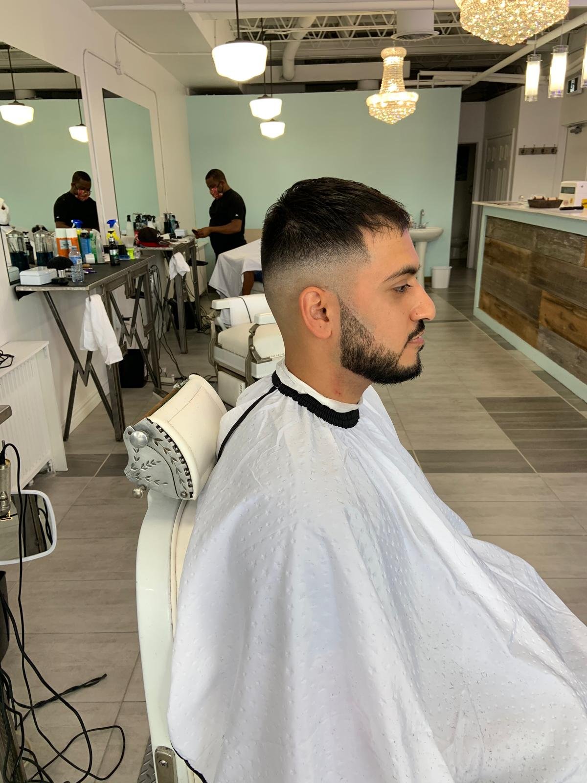 A man with a beard and short hair sits in a barber chair in a modern barbershop, facing to the right. The barber shop has a light blue wall, mirrors, and various hair and grooming products on the counter in the background.
