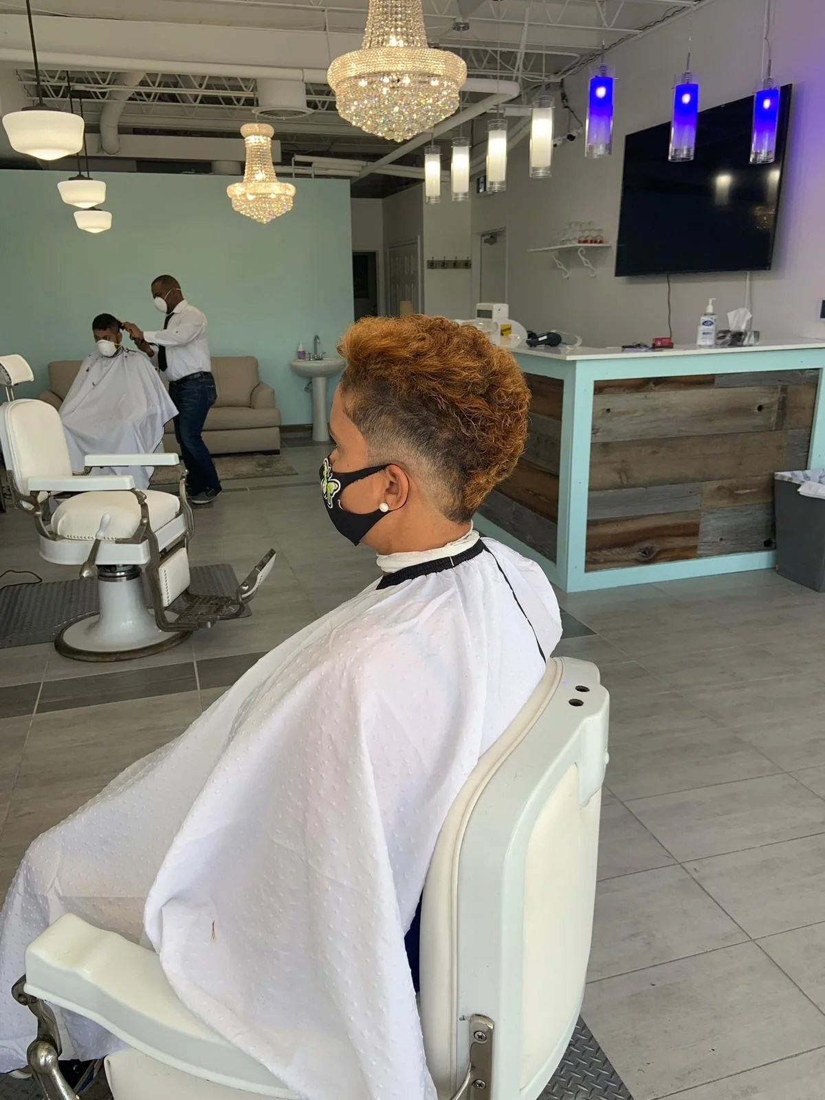 A woman having her hair cut at a barbershop, seated in a barber chair with a white cape and face mask, with a stylist in the background also wearing a face mask, working on another customer, in a modern salon with chandeliers and mounted TV screens.