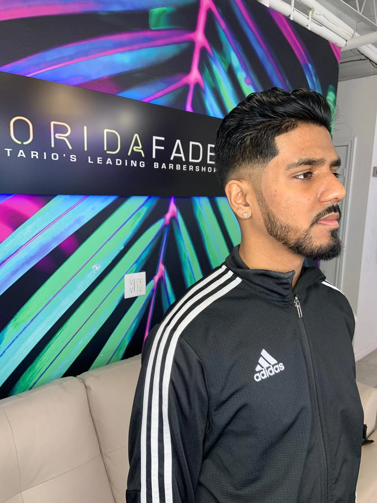 A man with dark hair and beard wearing a black Adidas jacket with white stripes, standing in front of a colorful neon-themed wall and a sign that reads "Orida Fade, Tario's Leading Barbershop."