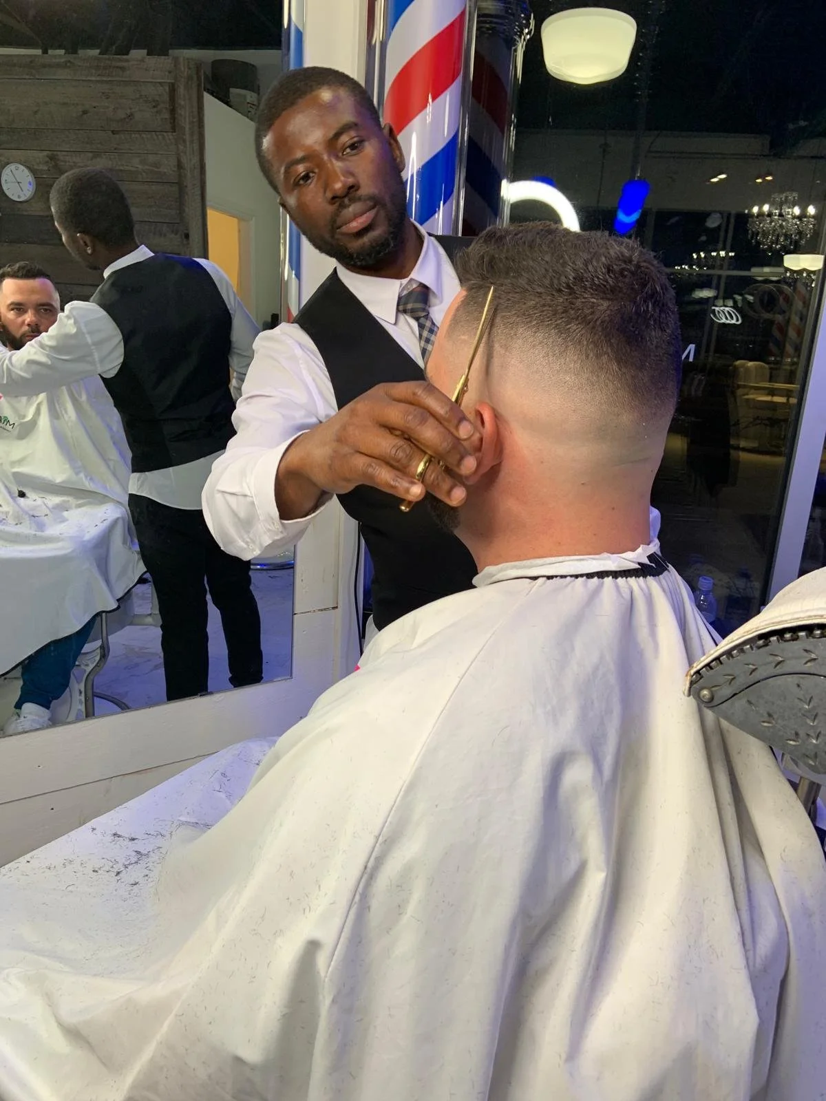 Barber cutting a customer's hair in a barbershop, with a barber holding scissors, wearing a white shirt, black vest, and tie. The customer is seated, wearing a cape. There is a large mirror behind the barber and another barber working in the backgrou