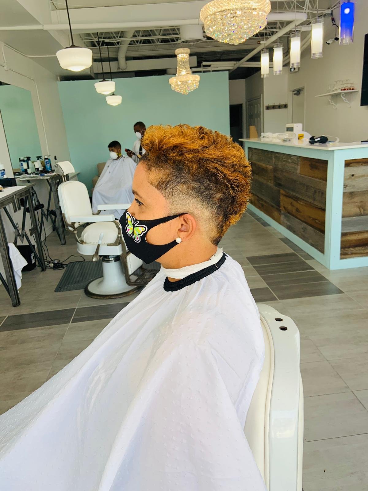 A woman with short curly hair styled with fade on the sides and back, wearing a butterfly face mask and pearl earrings, sits in a hair salon chair, preparing for a haircut or styling.