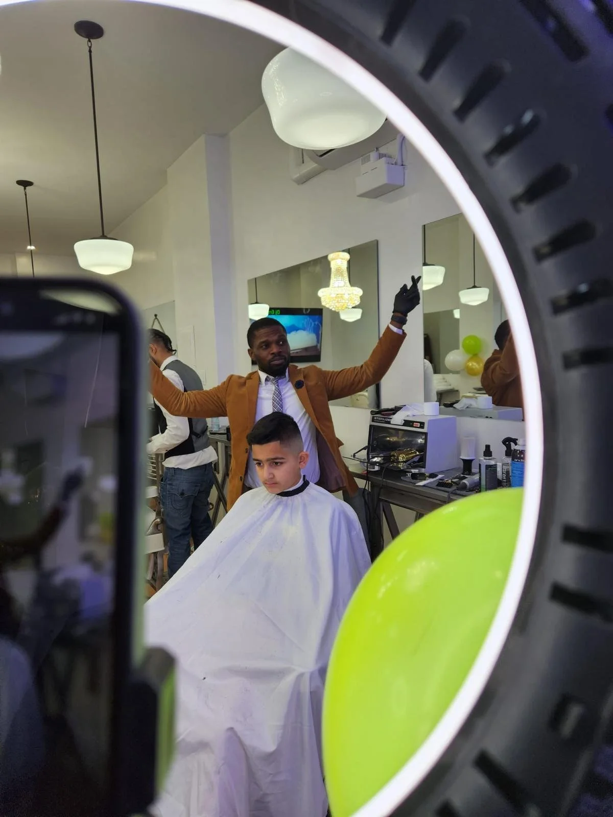 A man getting a haircut in a salon, seen through a mirror and a ring light, with a young boy in a barber cape sitting in the chair.