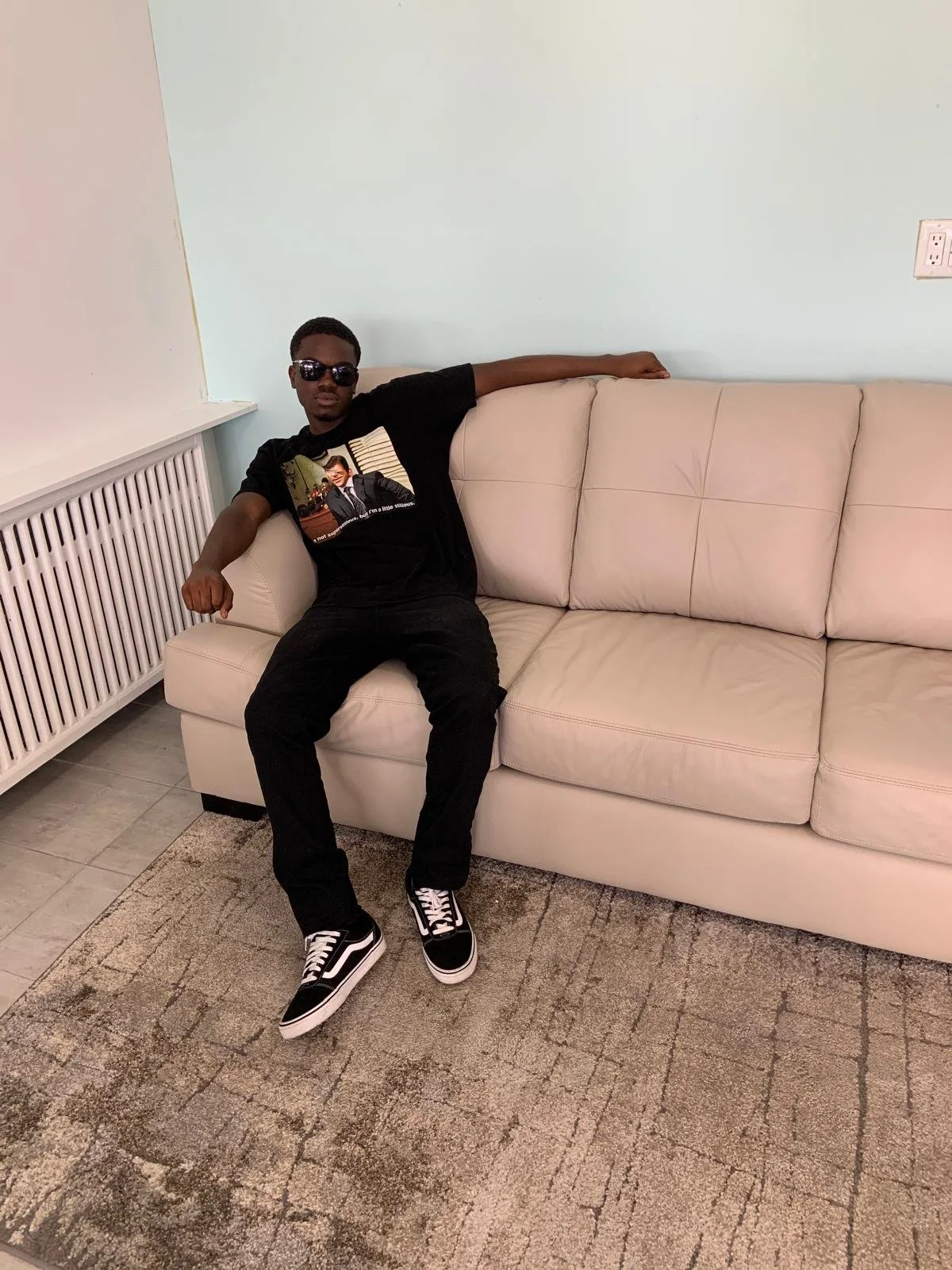 A young man wearing sunglasses, a black graphic t-shirt, black pants, and black and white Vans sneakers, sitting on a cream-colored sofa with his arm resting along the back of the sofa, in a room with light blue walls and a beige and gray patterned r