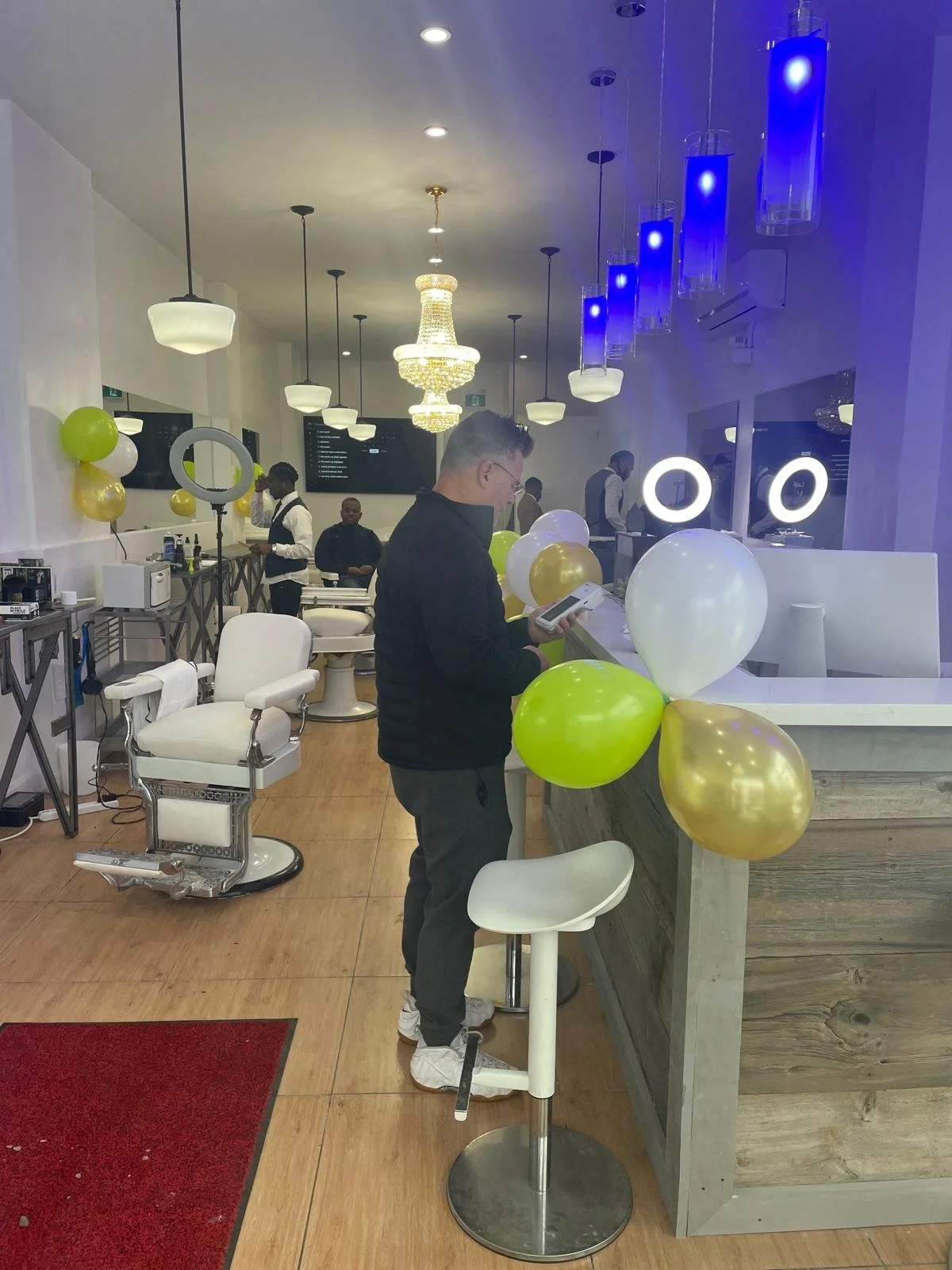People in a salon decorated with gold and white balloons, some getting haircuts or treatments, with elegant lighting and modern decor.