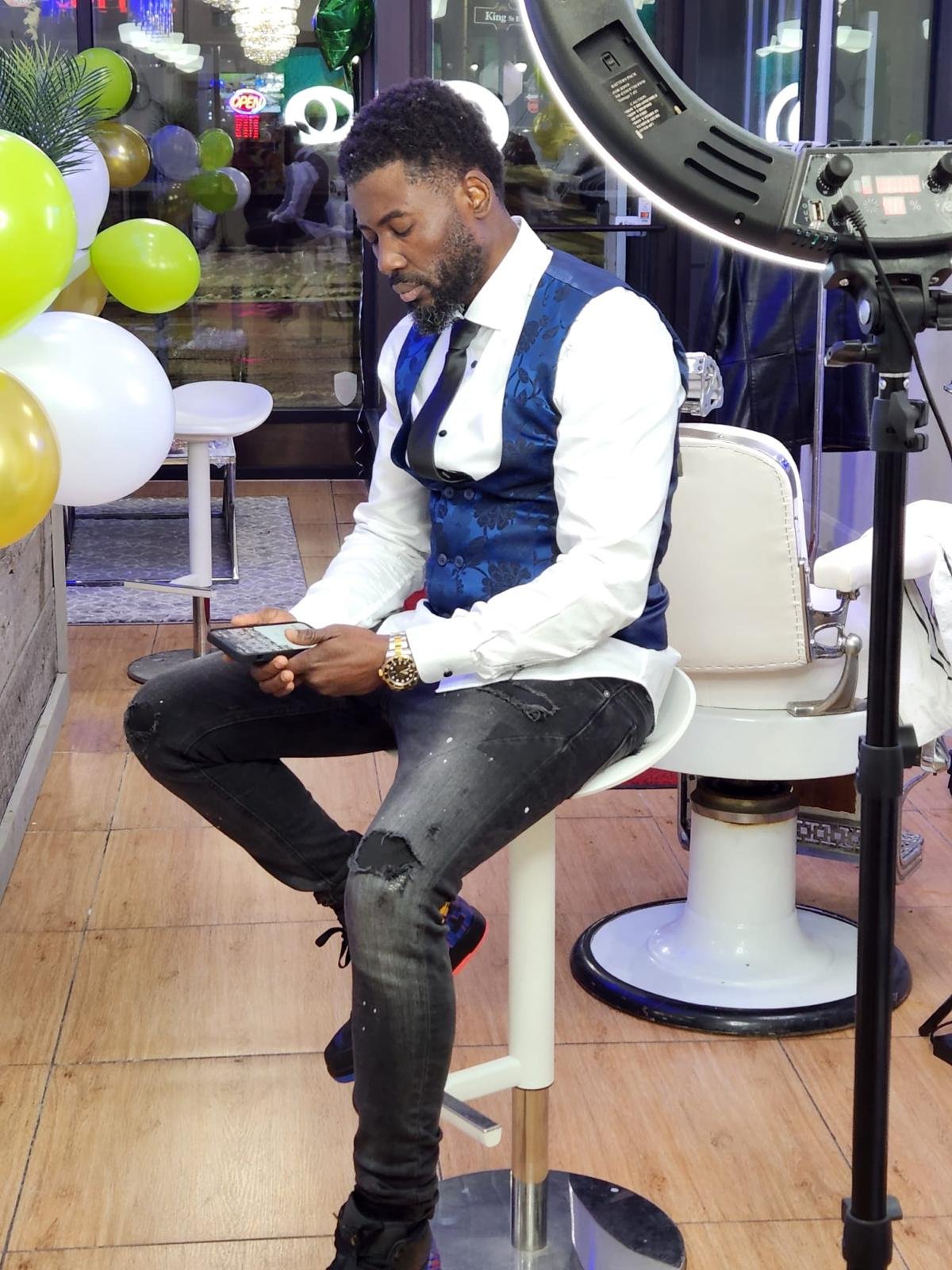 A man sitting on a barber's chair in a salon, looking at his phone. He is wearing a white shirt with a blue vest and black jeans, with a watch on his left wrist. There are balloons and a ring light setup around him, indicating a special event or phot
