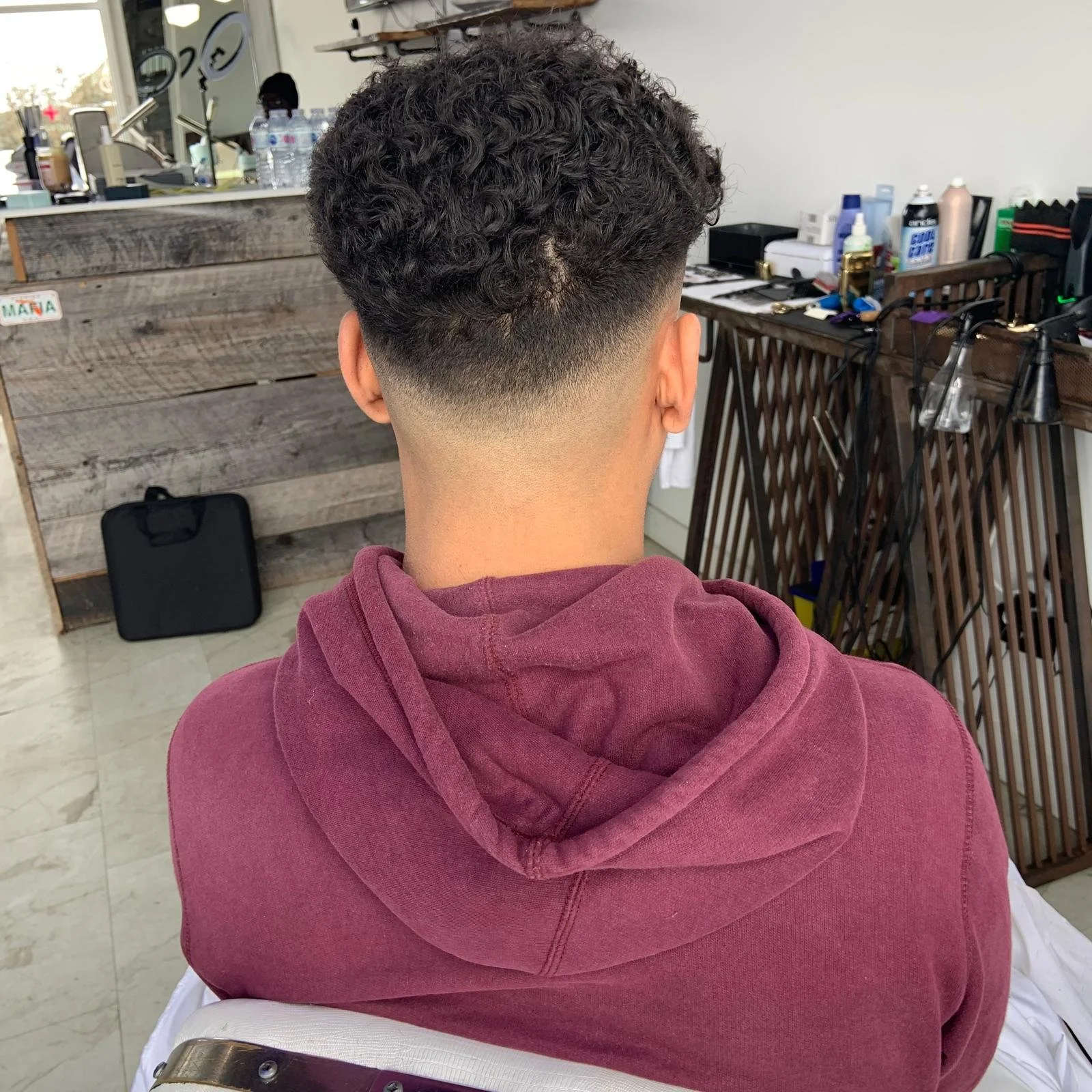 Back of a person's head with a fresh fade haircut, wearing a maroon hoodie, sitting in a barbershop.