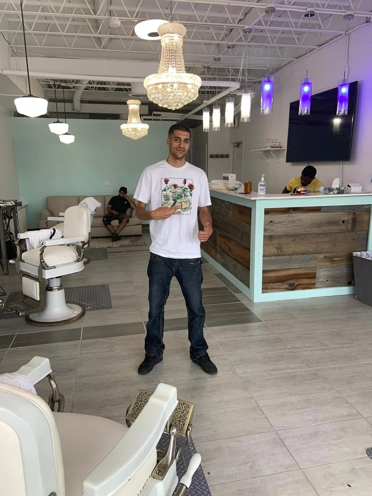 A young man standing in a modern salon, holding a card with an image of flowers and money, posing and smiling at the camera. The salon has chandeliers, pendant lights, and a reception/work area with two other employees working.