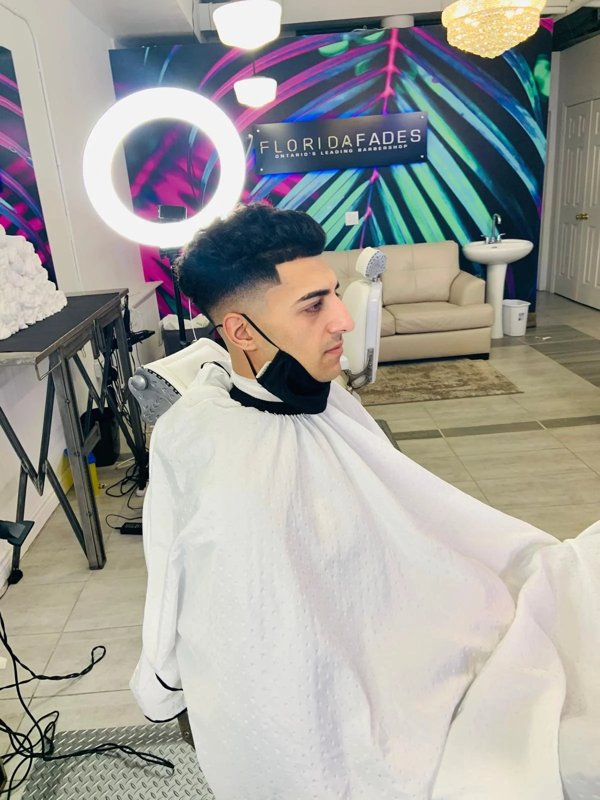 A young man with a fresh haircut sits in a barber chair at Florida Fades, a barbershop with vibrant tropical leaf patterned wallpaper and illuminated circular mirror. There is a beige sofa, sink, and mirror in the background, with colorful lighting.