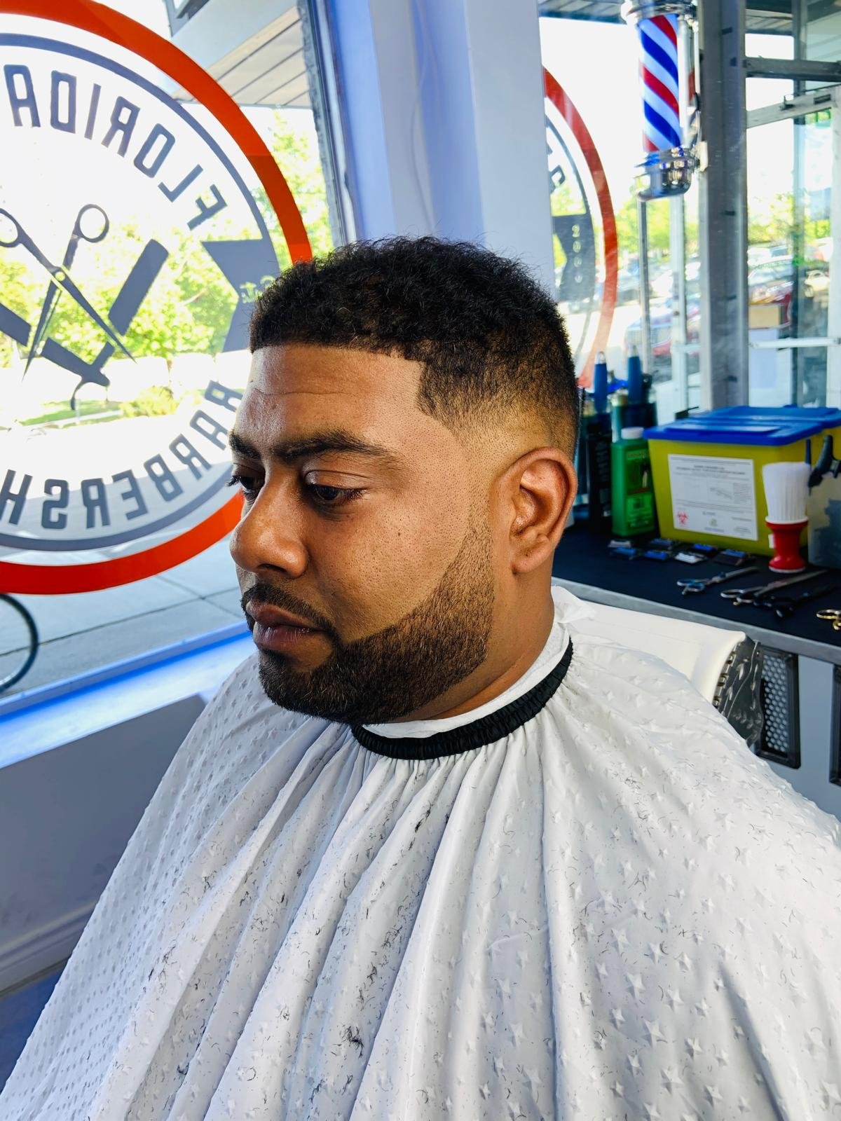 Man with a fresh haircut sitting in a barbershop chair, wearing a white barber cape, inside a barbershop with various tools and products in the background, and a glass window with a logo outside.